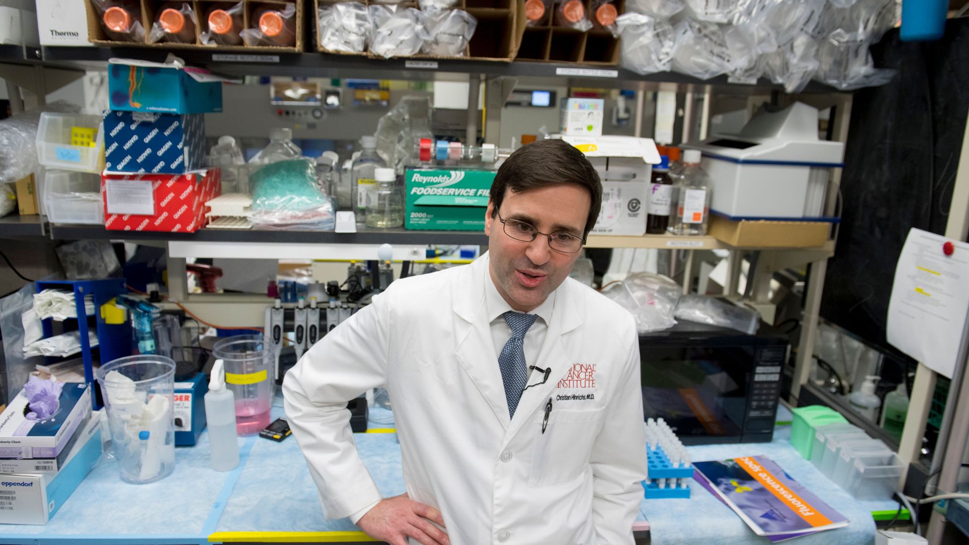 A researcher at the National Cancer Institute stands in a white lab coat in front of several shelves of medical supplies.