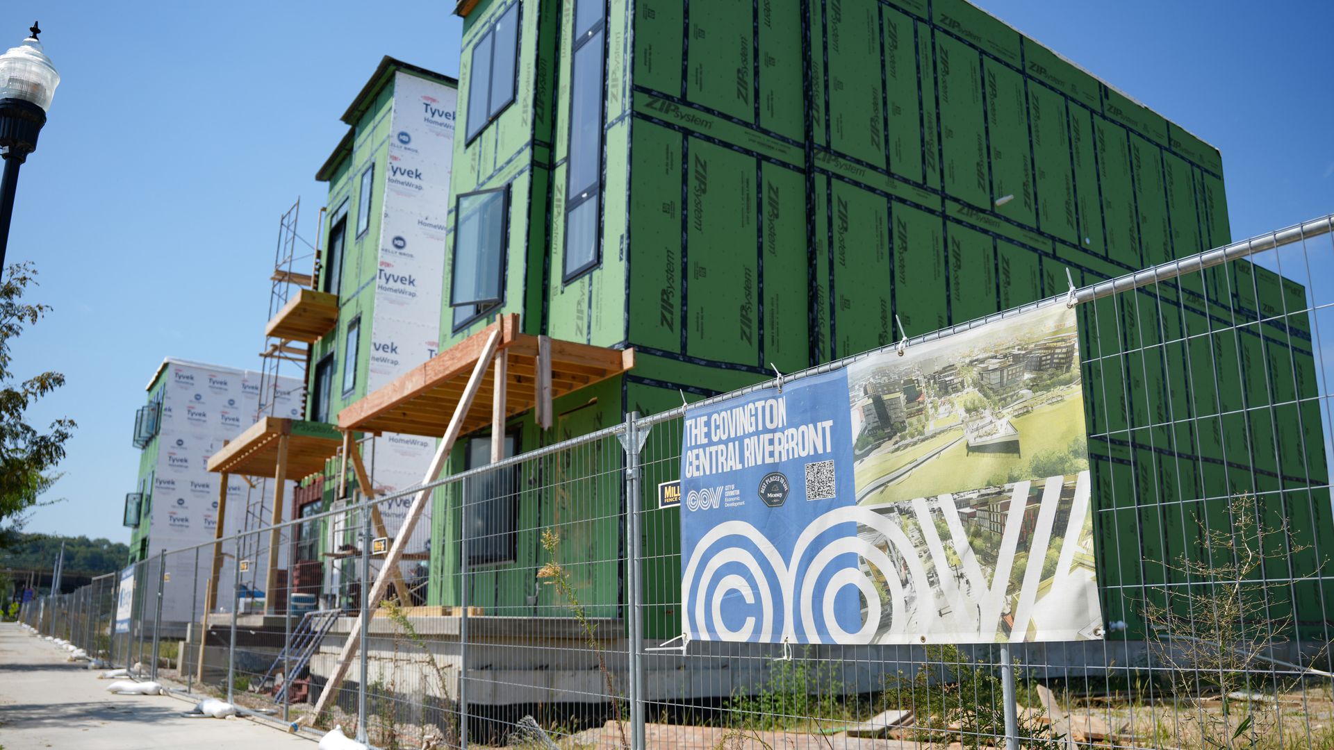 A construction site with a green-paneled building, black-framed windows, wooden scaffolding, white Tyvek wrap on the left, and a chain-link fence with a blue banner under a clear sky.