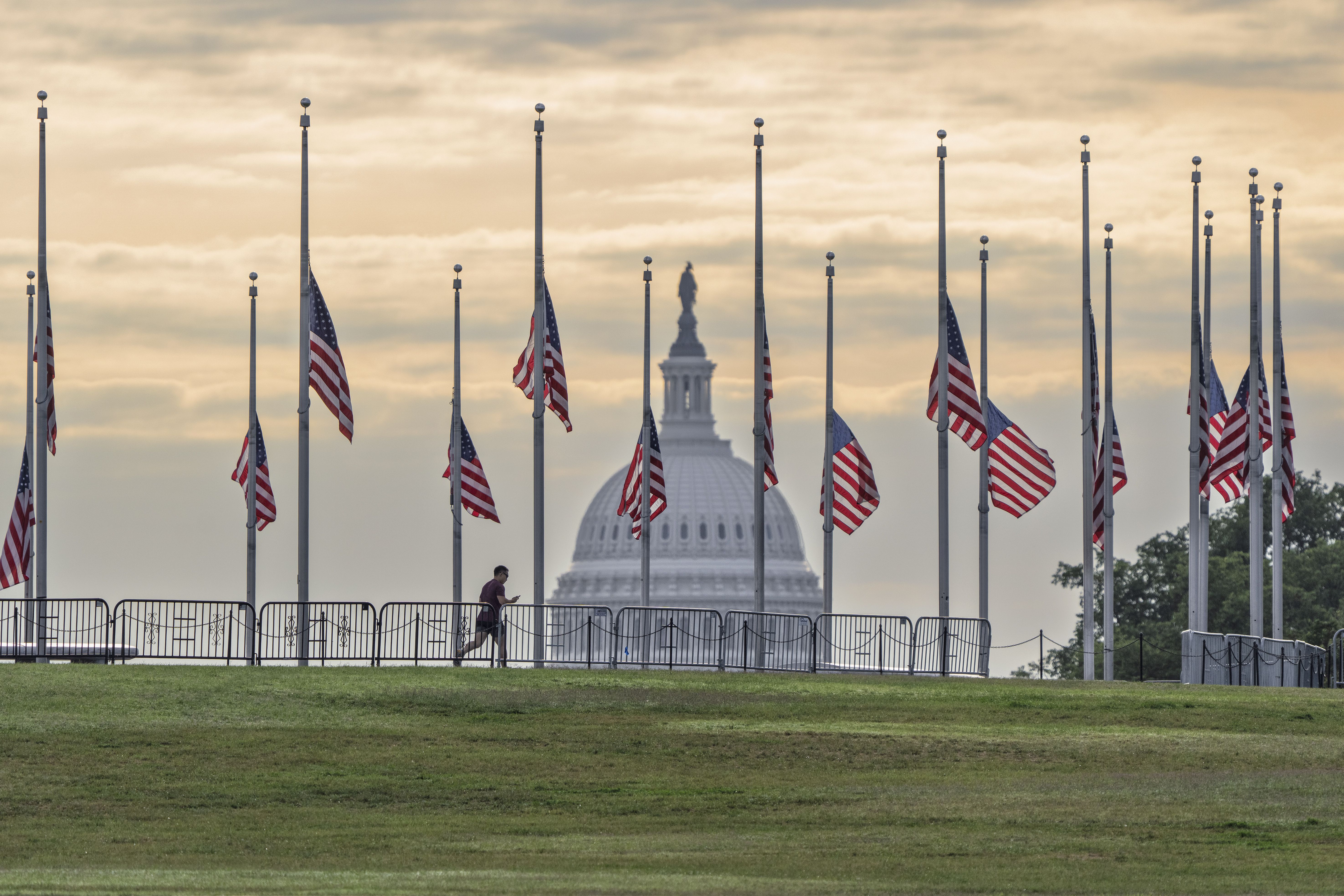View of the U.S. Capitol dome at sunset behind rows of American flags flying at half-staff with a runner passing by on green grass in the foreground.