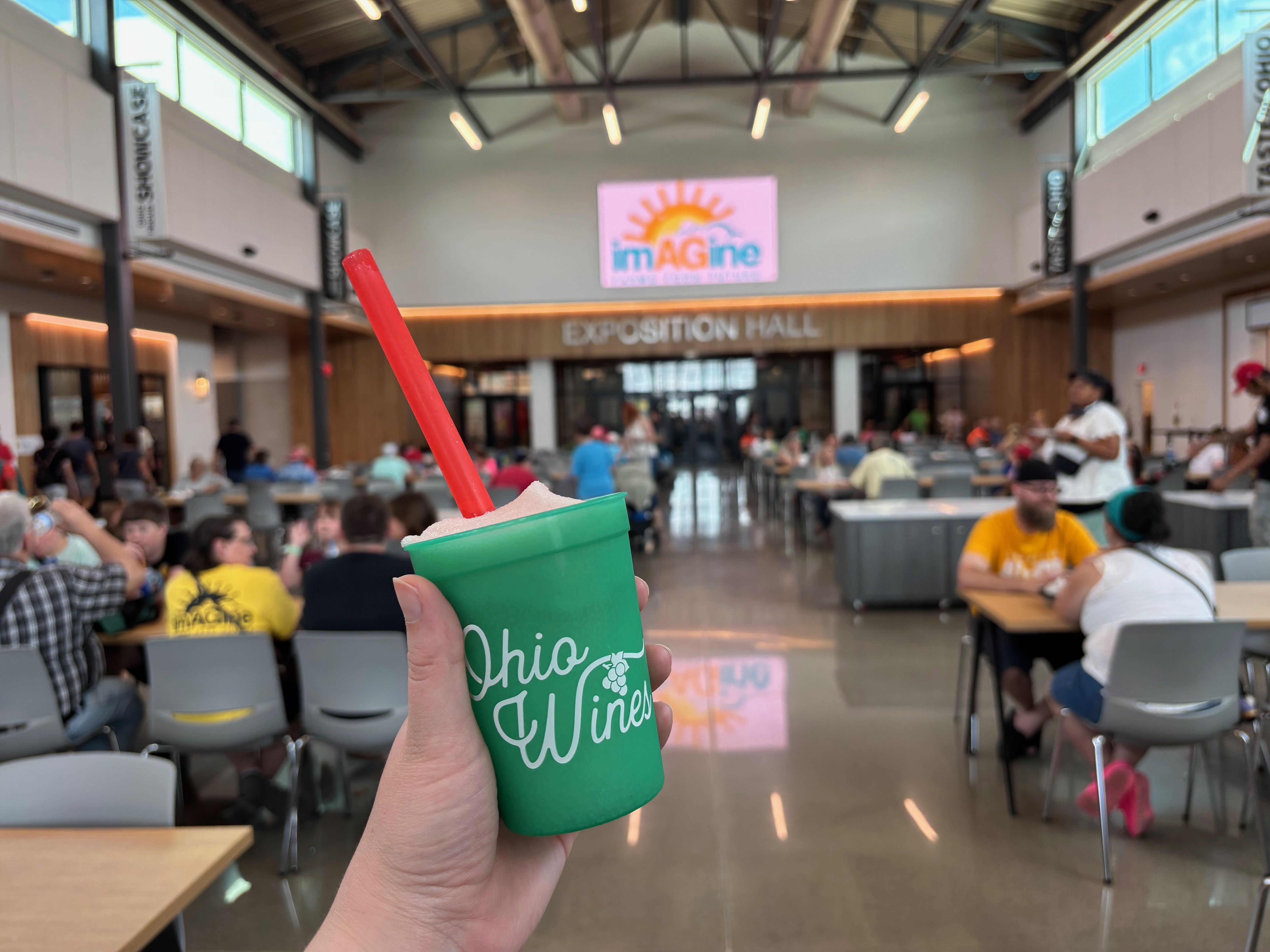 Hand holding a green cup with a red straw in a busy expo hall; blurred crowd at tables, a large "EXPOSITION HALL" sign, and a colorful "imAGine" logo on the wall.