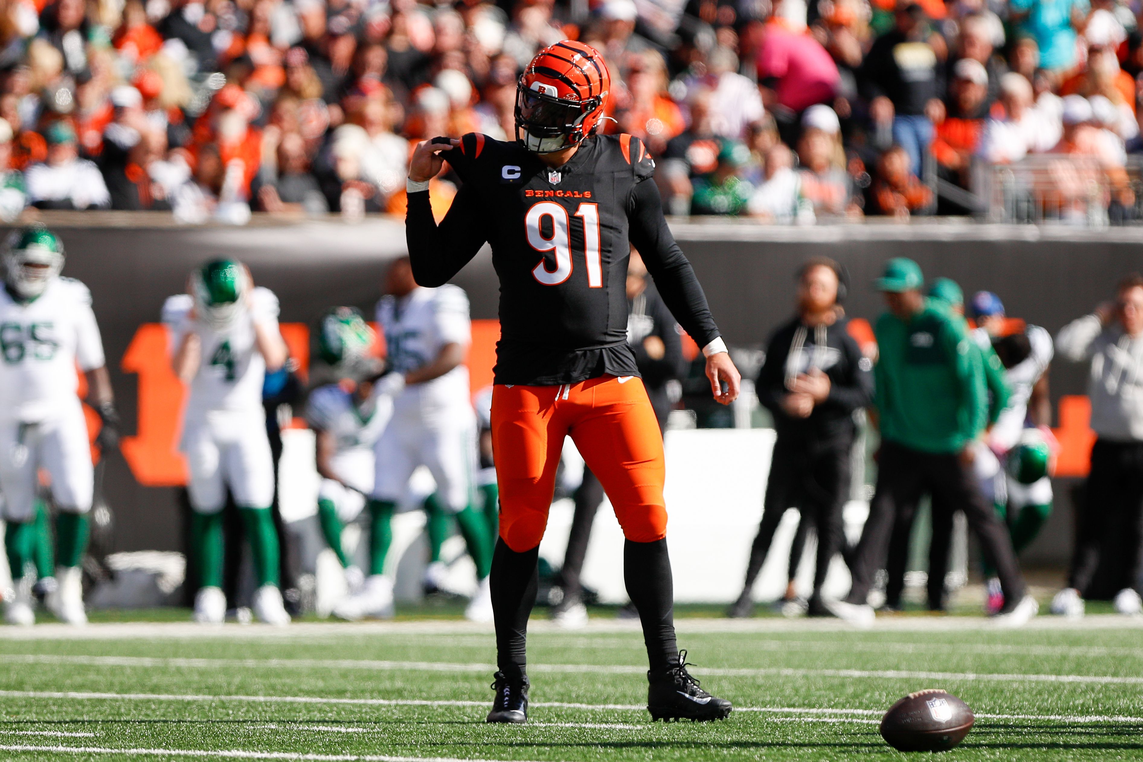 Cincinnati Bengals player #91 in black and orange uniform on football field with ball on ground, opposing team in white and green in background, stadium crowd visible.