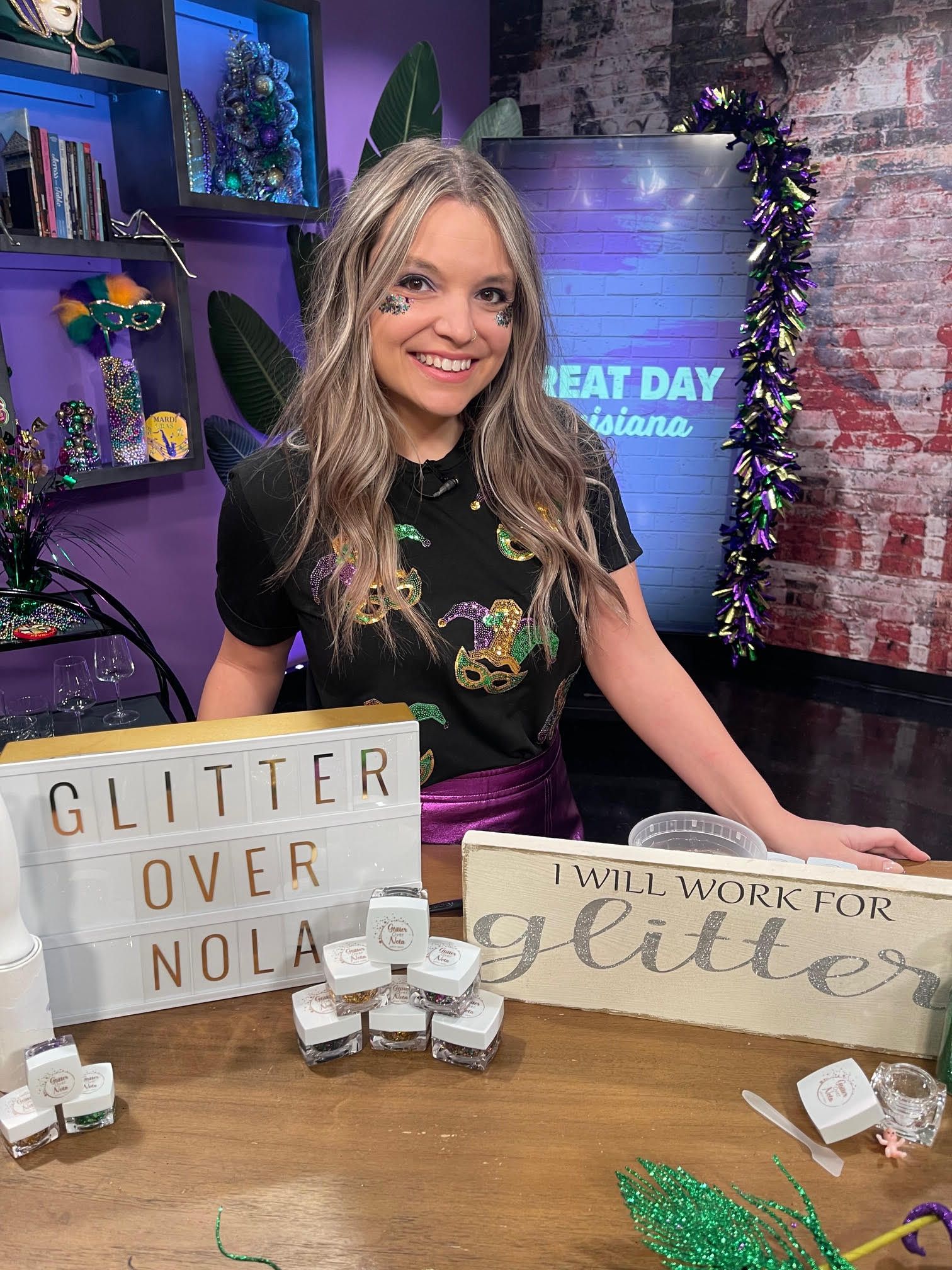 Smiling woman with glittery makeup on cheeks stands behind a table with glitter containers, signs reading "GLITTER OVER NOLA" and "I WILL WORK FOR glitter," in a colorful Mardi Gras-themed room.