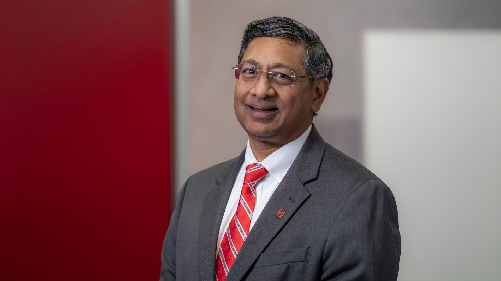 A headshot of Ravi V. Bellamkonda in a gray suit, white shirt, and red striped tie stands in an office with a red wall on the left and a light-gray wall behind, wearing glasses.