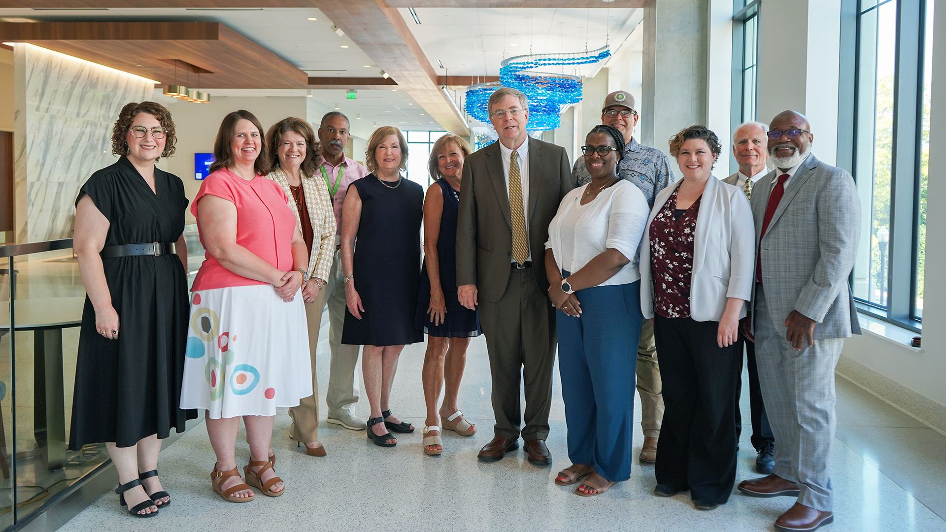 Group of professionals posing in a bright, modern lobby with a marble wall, wooden ceiling beams, and a blue hanging sculpture near large windows.
