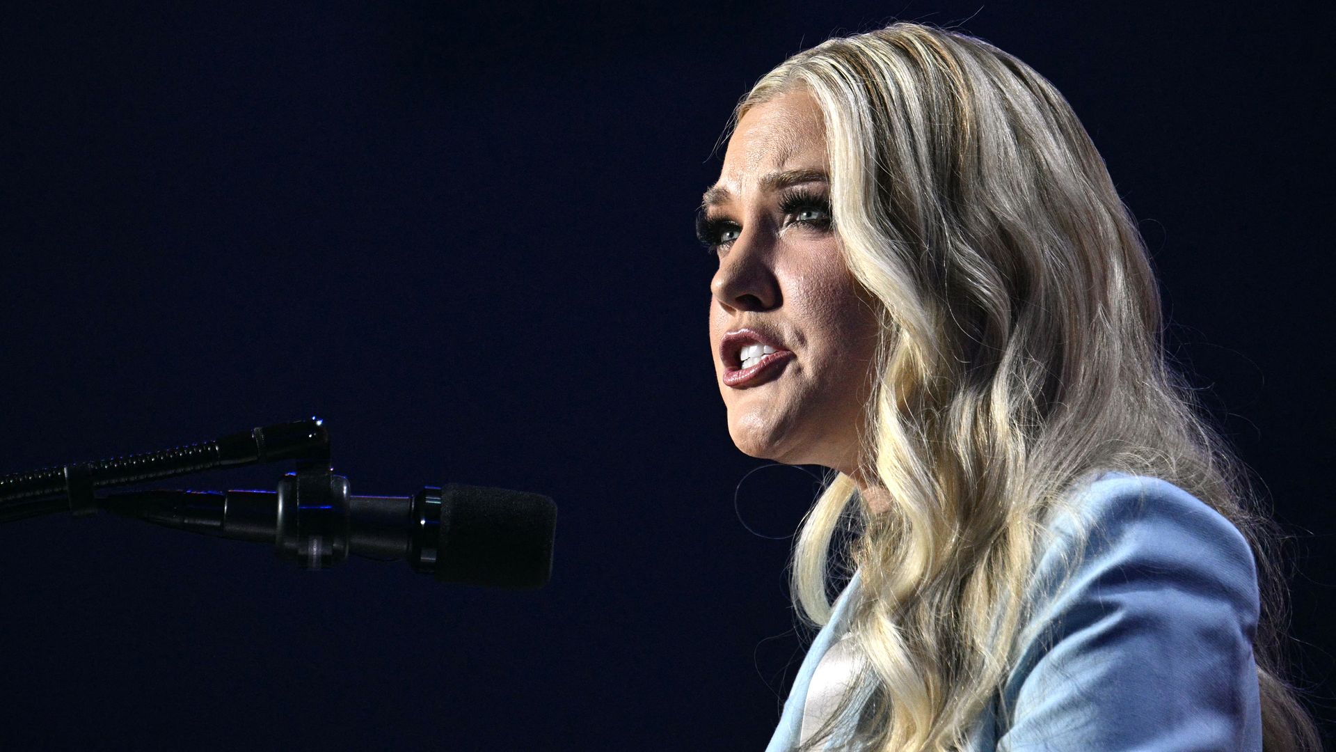 Erika Kirk, in a baby blue suit, stares out with the background behind her completely black. A microphone is seen in front of her.