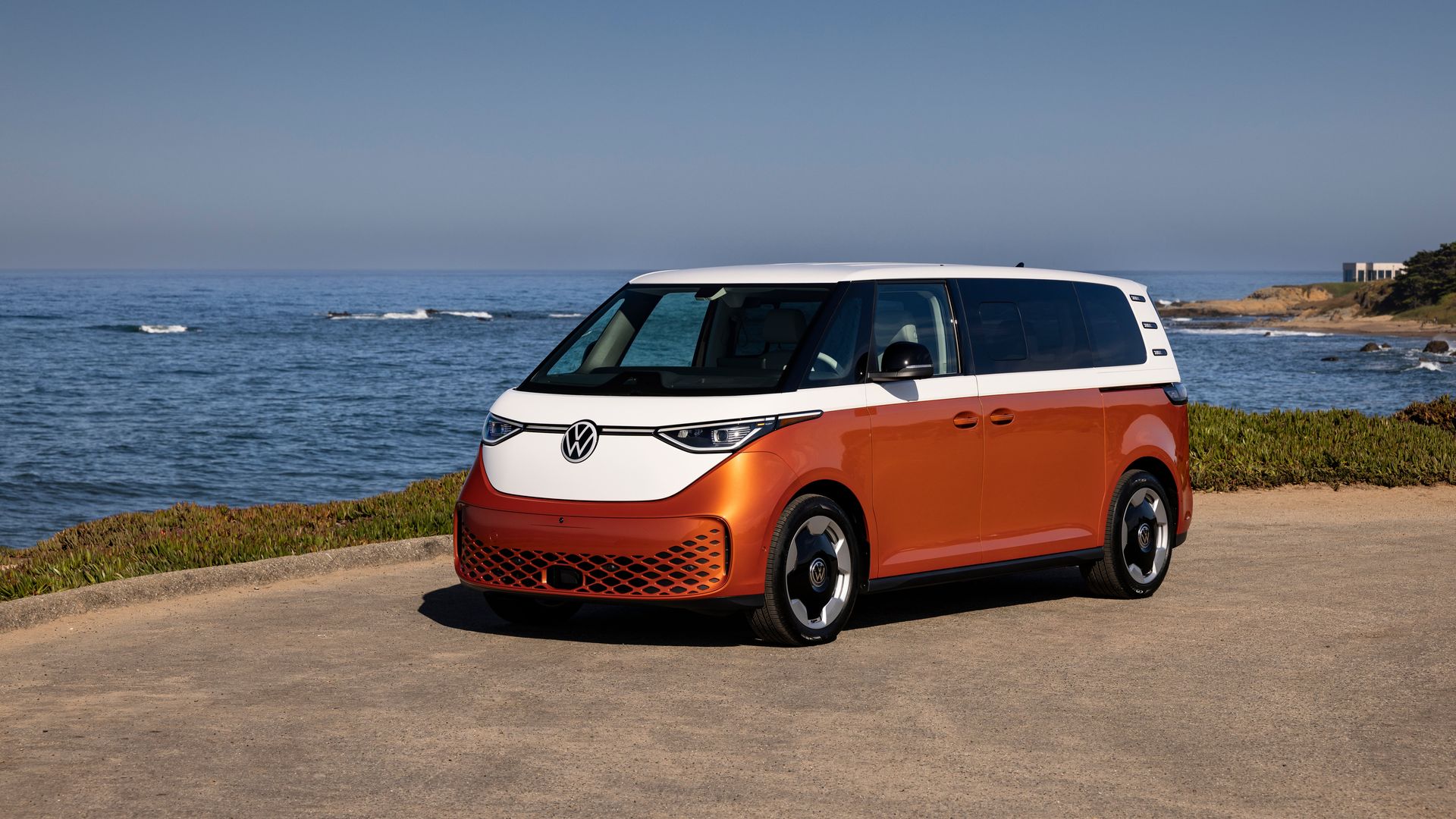 Orange and white Volkswagen electric van parked on a coastal road with ocean and rocks in the background under a clear blue sky.