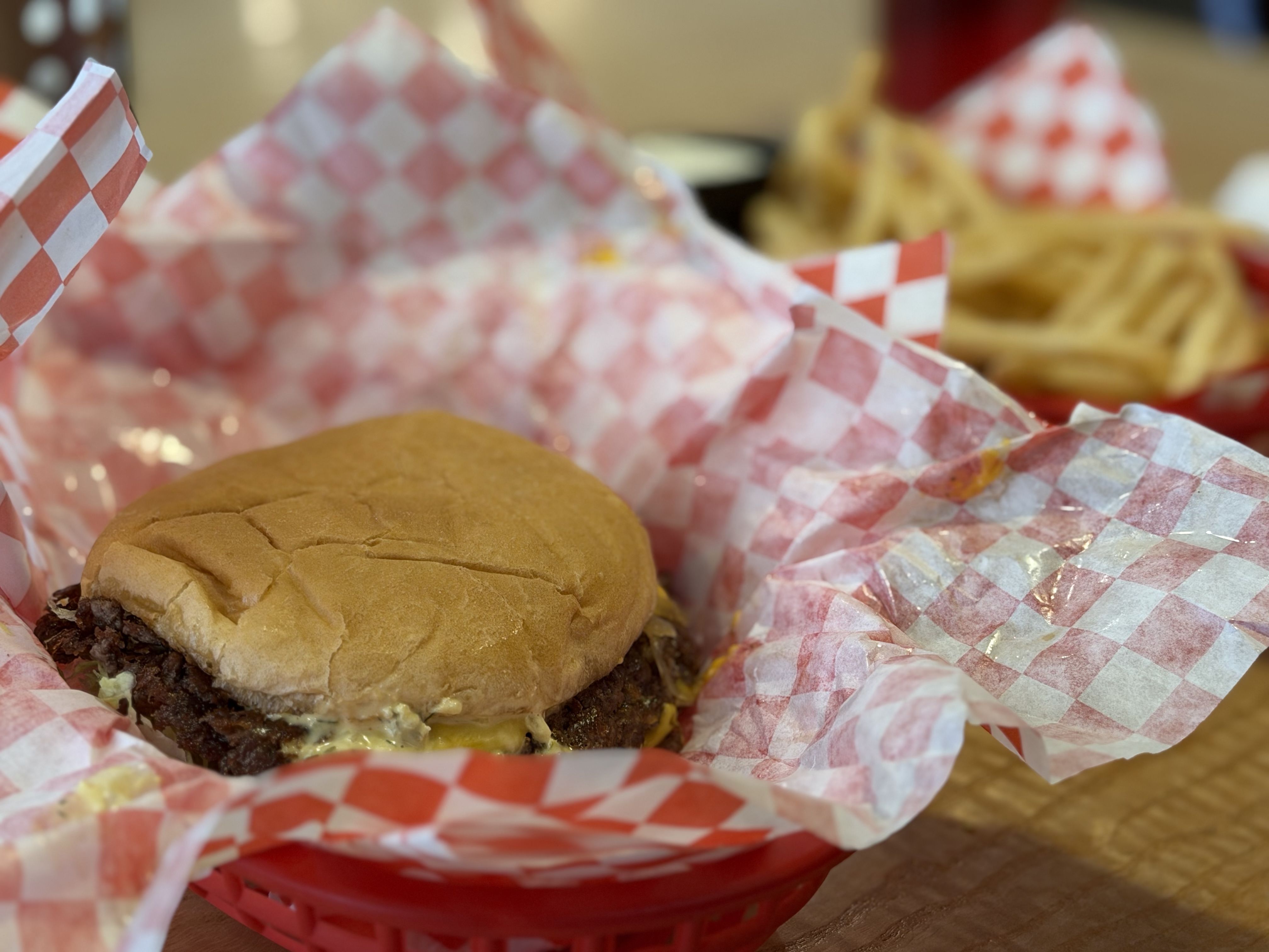 Close-up of a cheeseburger wrapped in red-and-white checkered paper, featuring a beef patty with melted cheese and a soft bun, with fries visible in the blurred background.