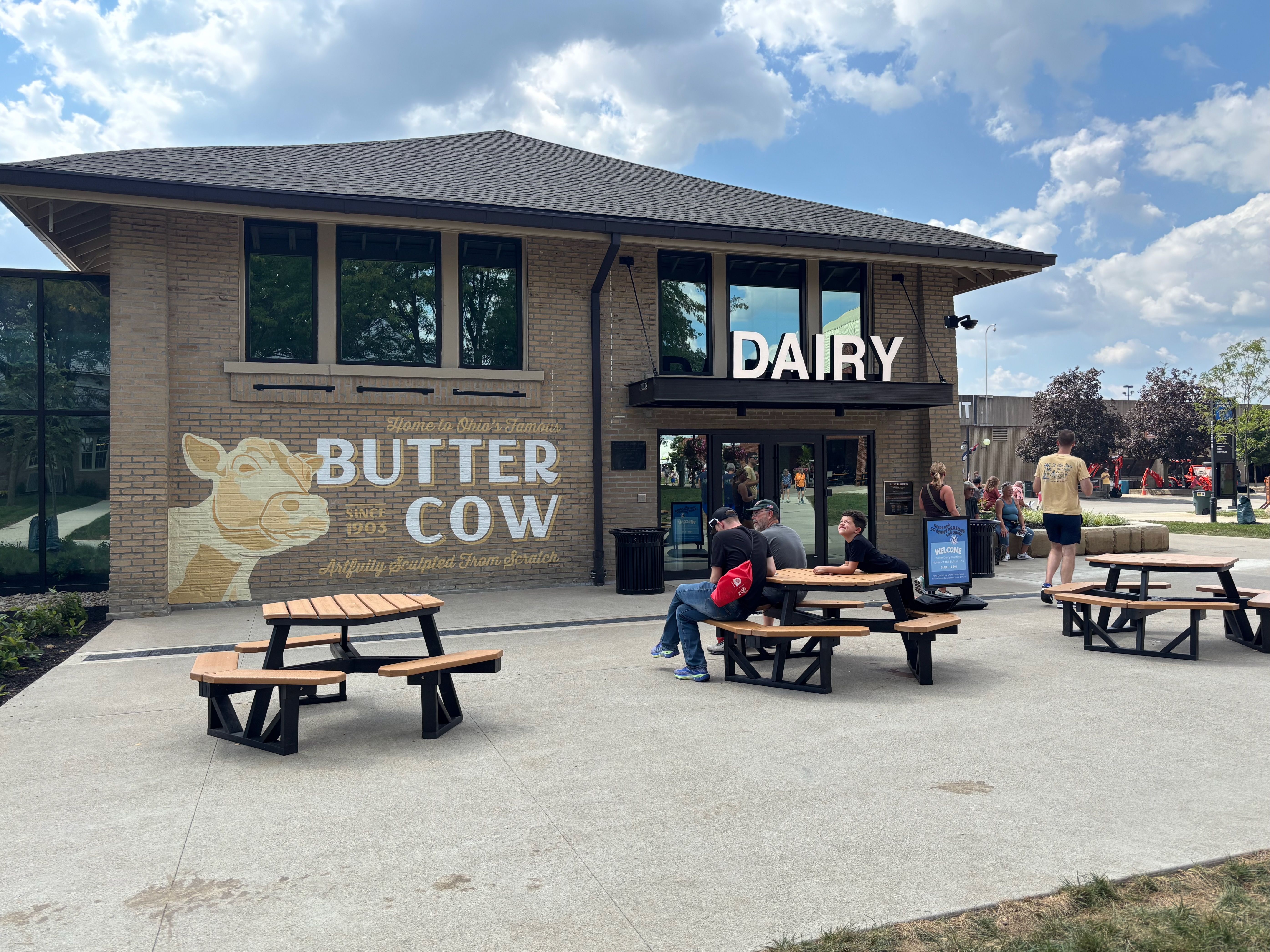 Brick dairy storefront with a large white DAIRY sign over glass doors. Wall mural reads BUTTER COW with a cow image. Outdoor picnic tables and people sit outside under a blue, cloudy sky.