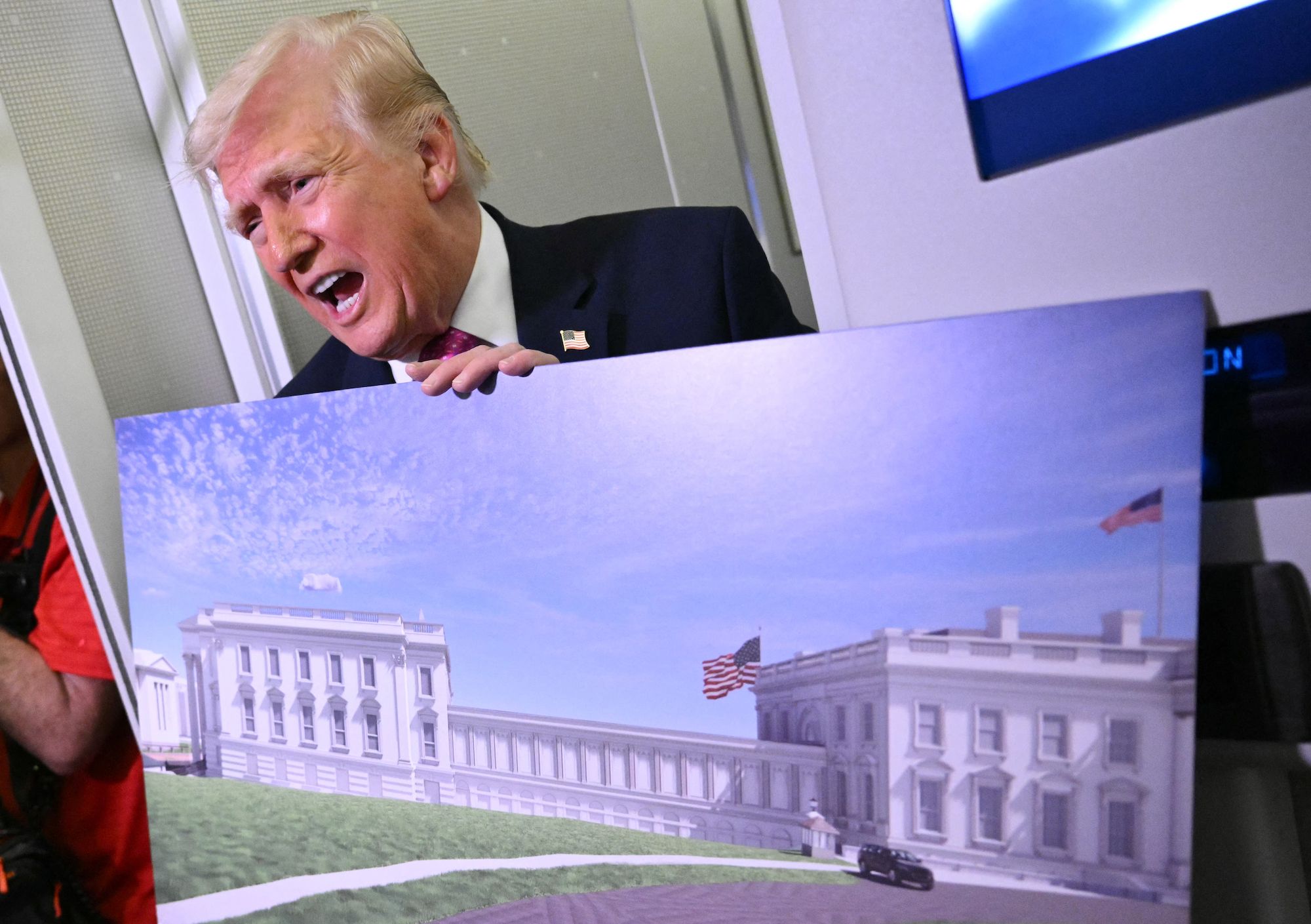 Donald Trump in a dark suit and red tie leans over, holding a large print of a white neoclassical government building with American flags, set against a blue sky.