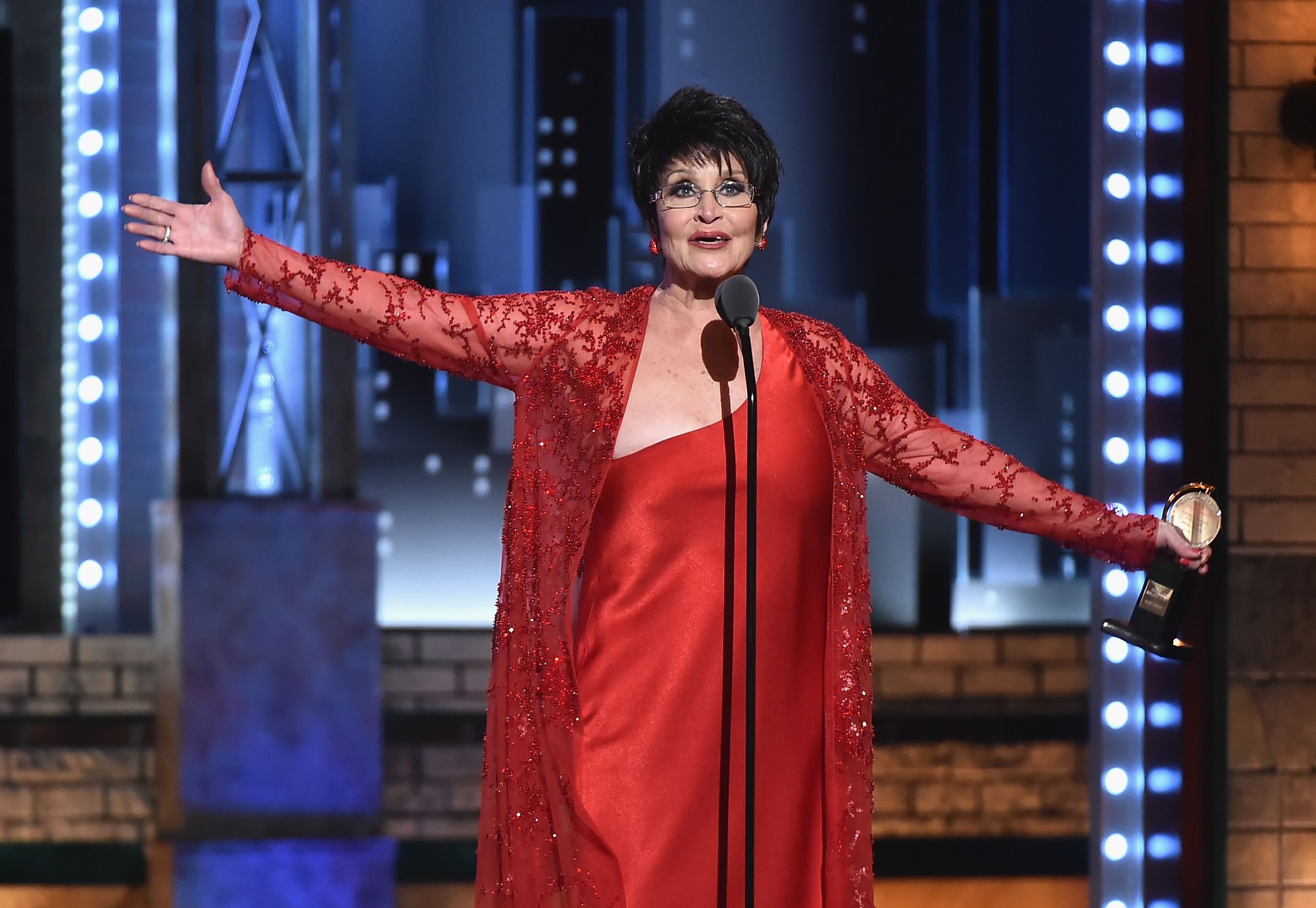Chita Rivera wearing all red stands with her arms out in front of a microphone with a stand at the Tony awards