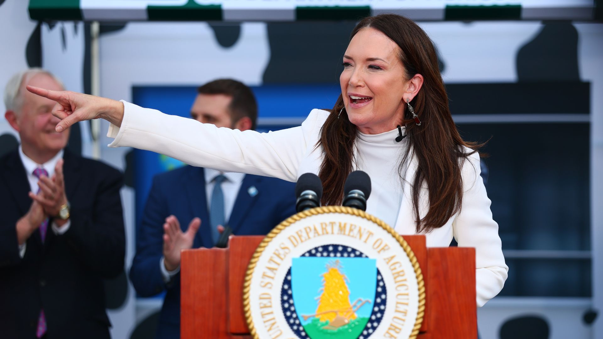 A woman in a white outfit speaks at a podium with the United States Department of Agriculture seal, pointing to her left while two men in suits clap behind her.