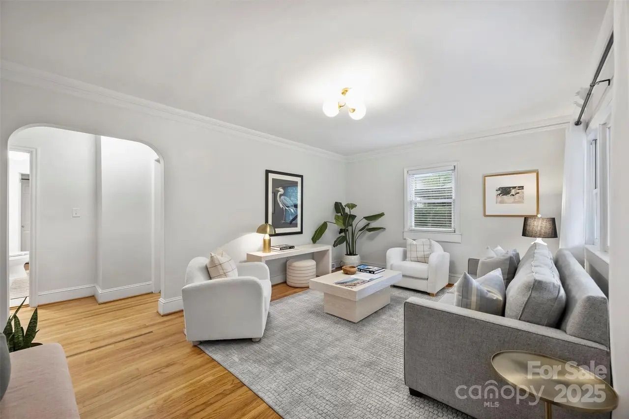 Bright living room with white walls, gray sofa, two white armchairs, light wood floor, a light-colored rug, coffee table, green plant, and wall art featuring a bird.