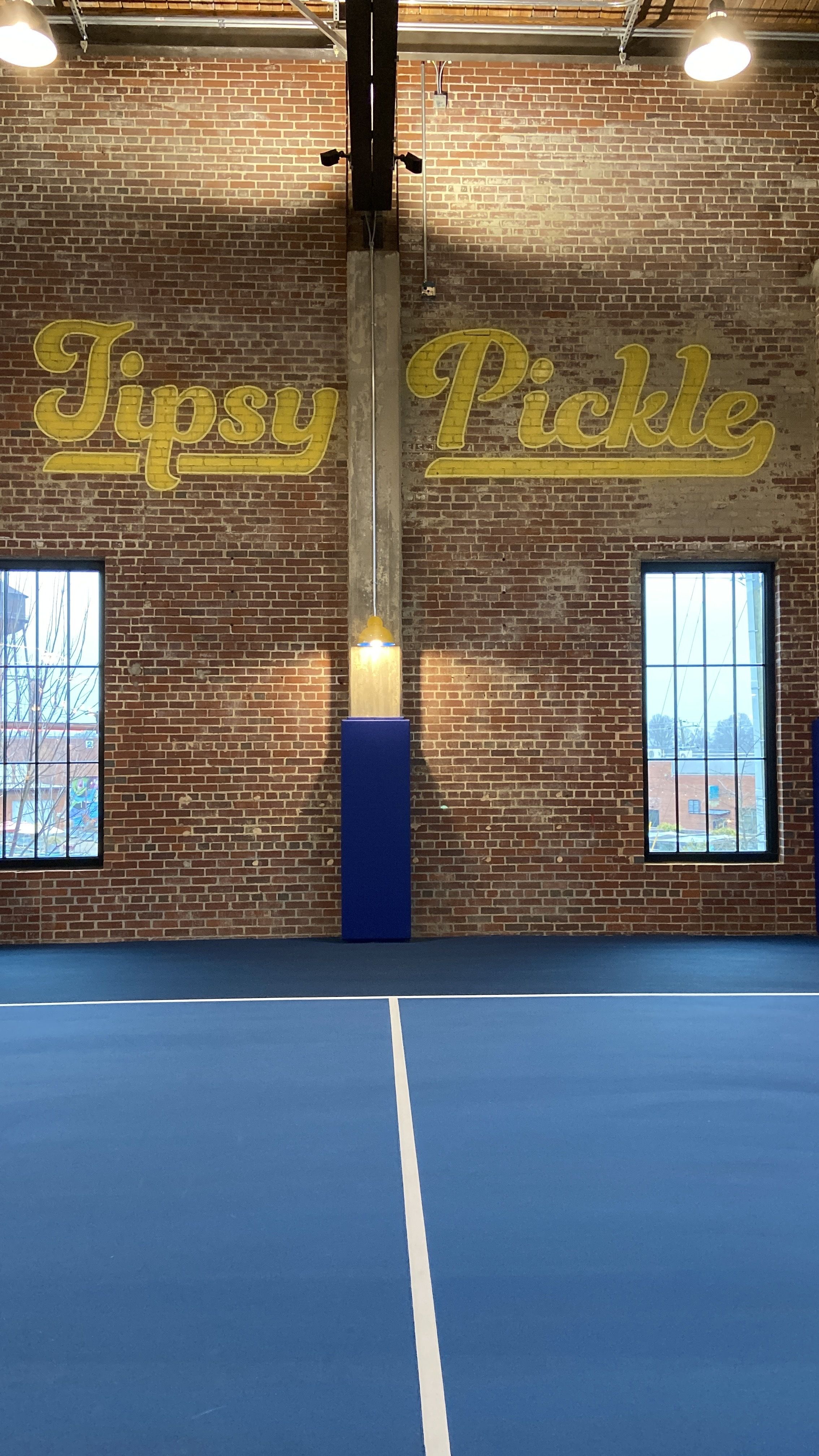 "Tipsy Pickle" painted in yellow on a brick wall above a blue pickleball court. 