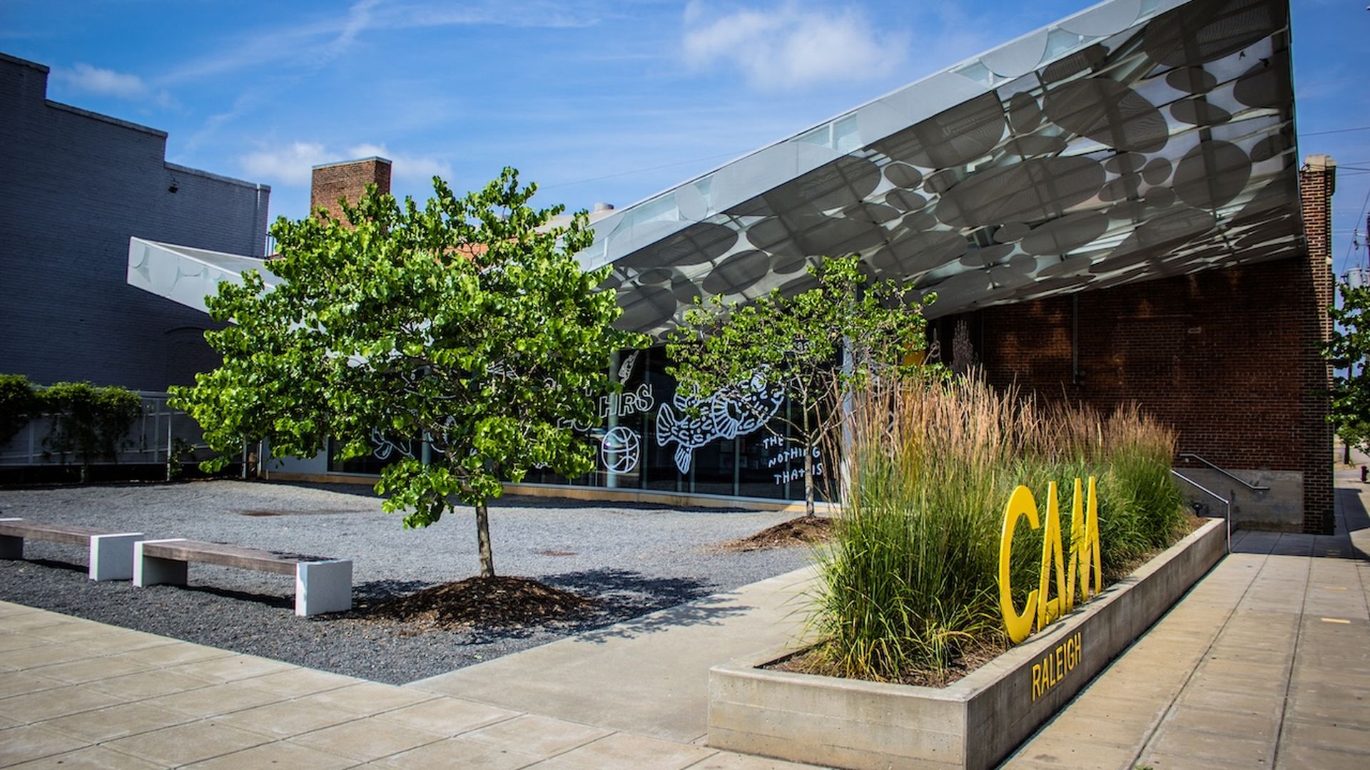 Exterior of a modern campus building with a curved glass canopy patterned with circles. A green tree sits in gravel beside a raised planter with large yellow CM letters and RALEIGH on the base.