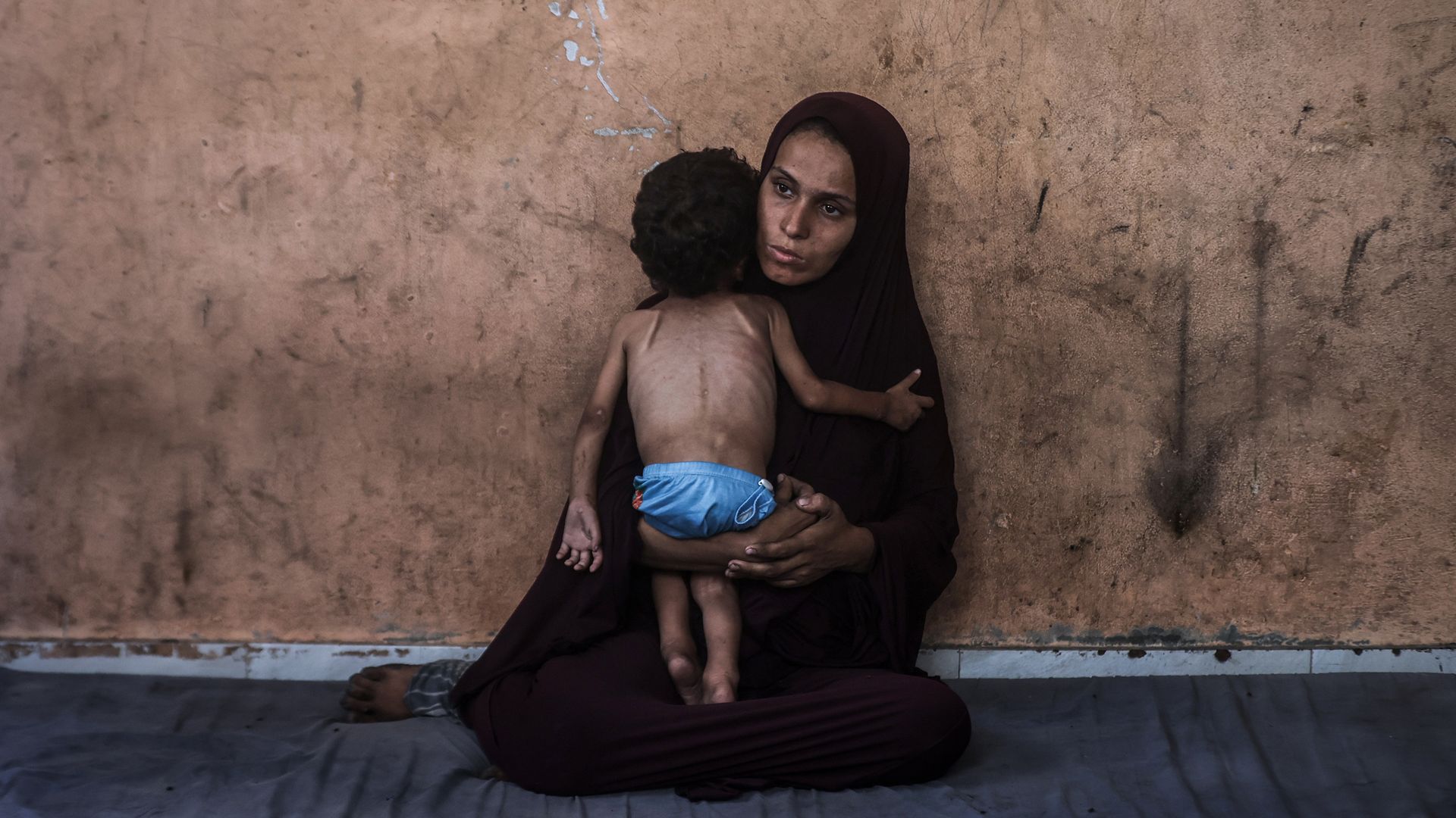 A woman in a dark hijab sits on the floor against a worn, brown wall, holding a small, thin child wearing only blue shorts who clings to her, conveying a sense of hardship and care.