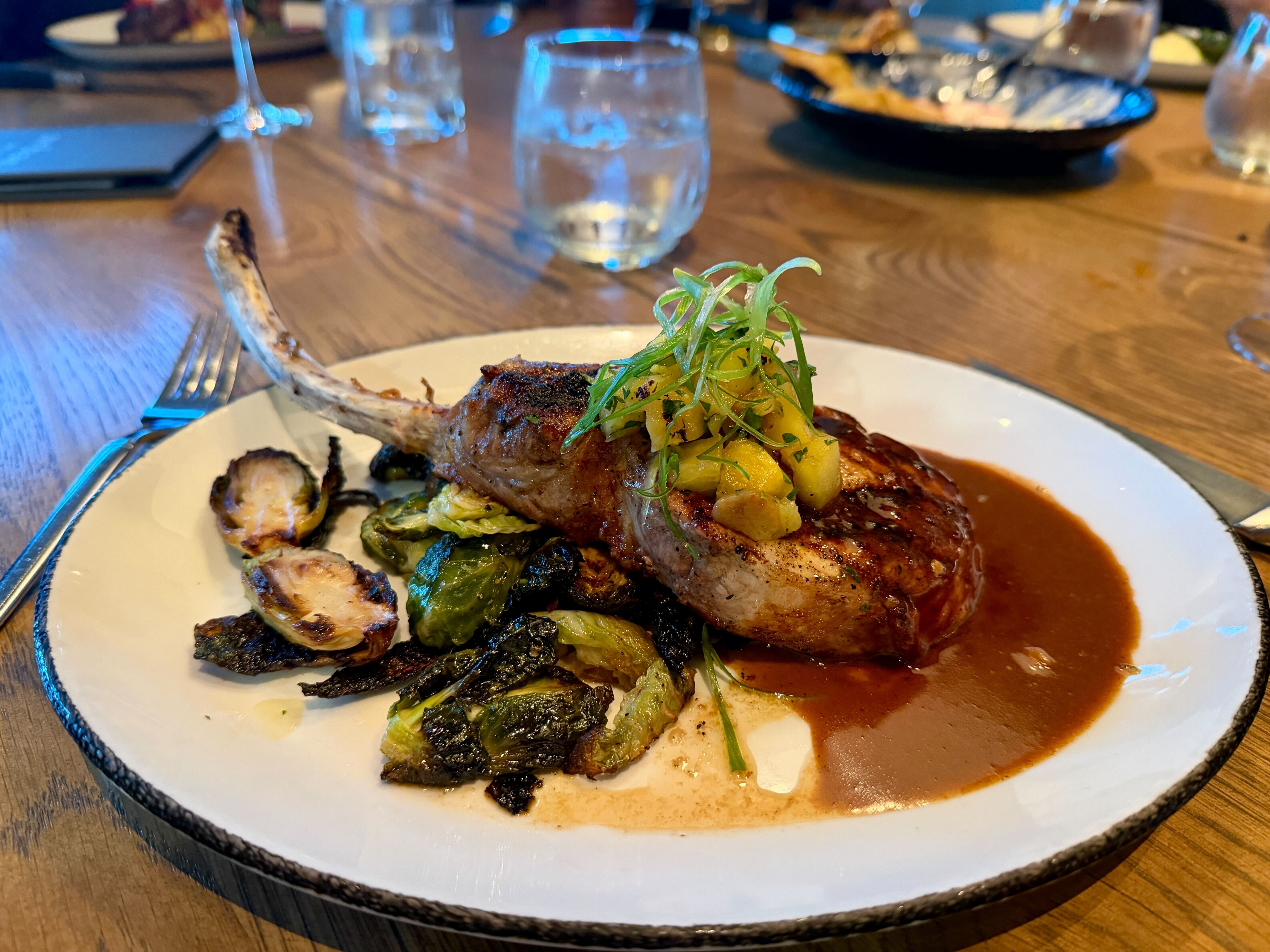 Plate with a roasted bone-in chop, brown sauce, and a yellow fruit relish topped with fresh green herb sprouts; roasted greens and vegetables on the side, a fork nearby on a wooden table.