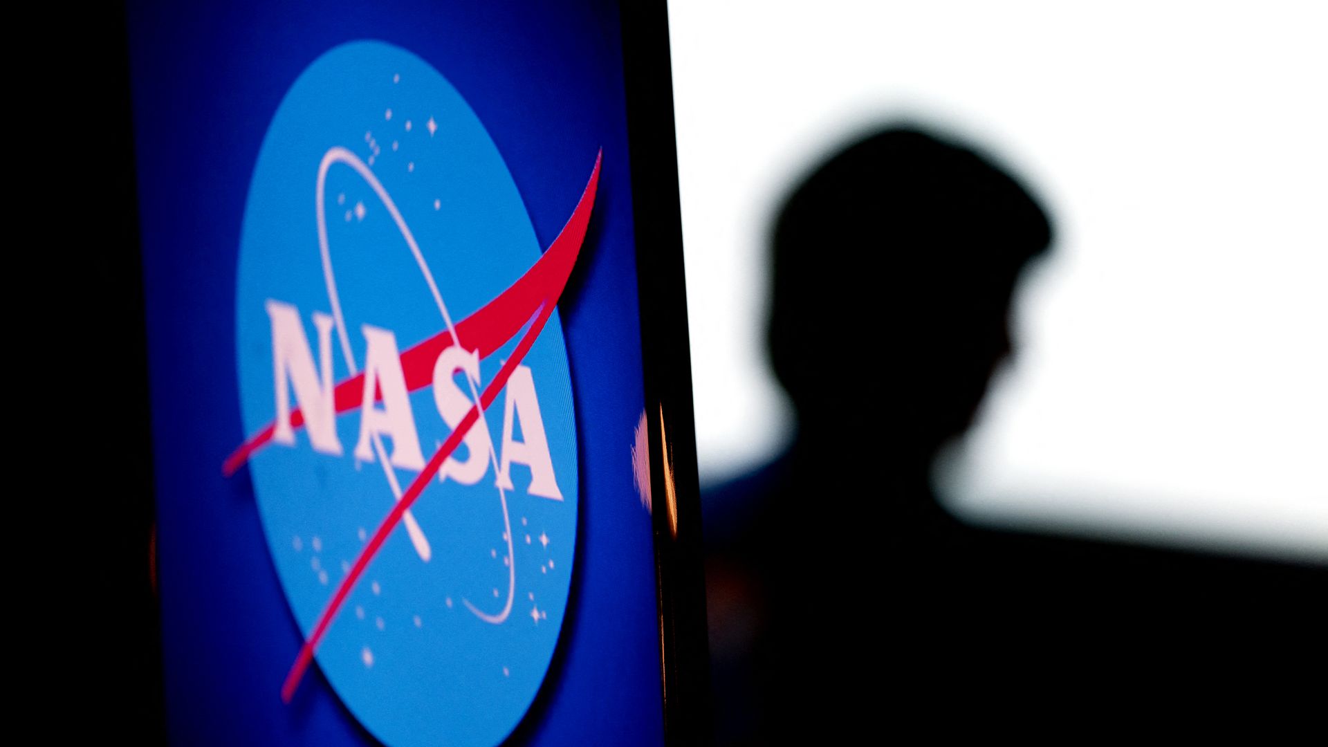 Close-up of the blue NASA logo with a red swoosh on the left, and a dark silhouetted person on a white background on the right.