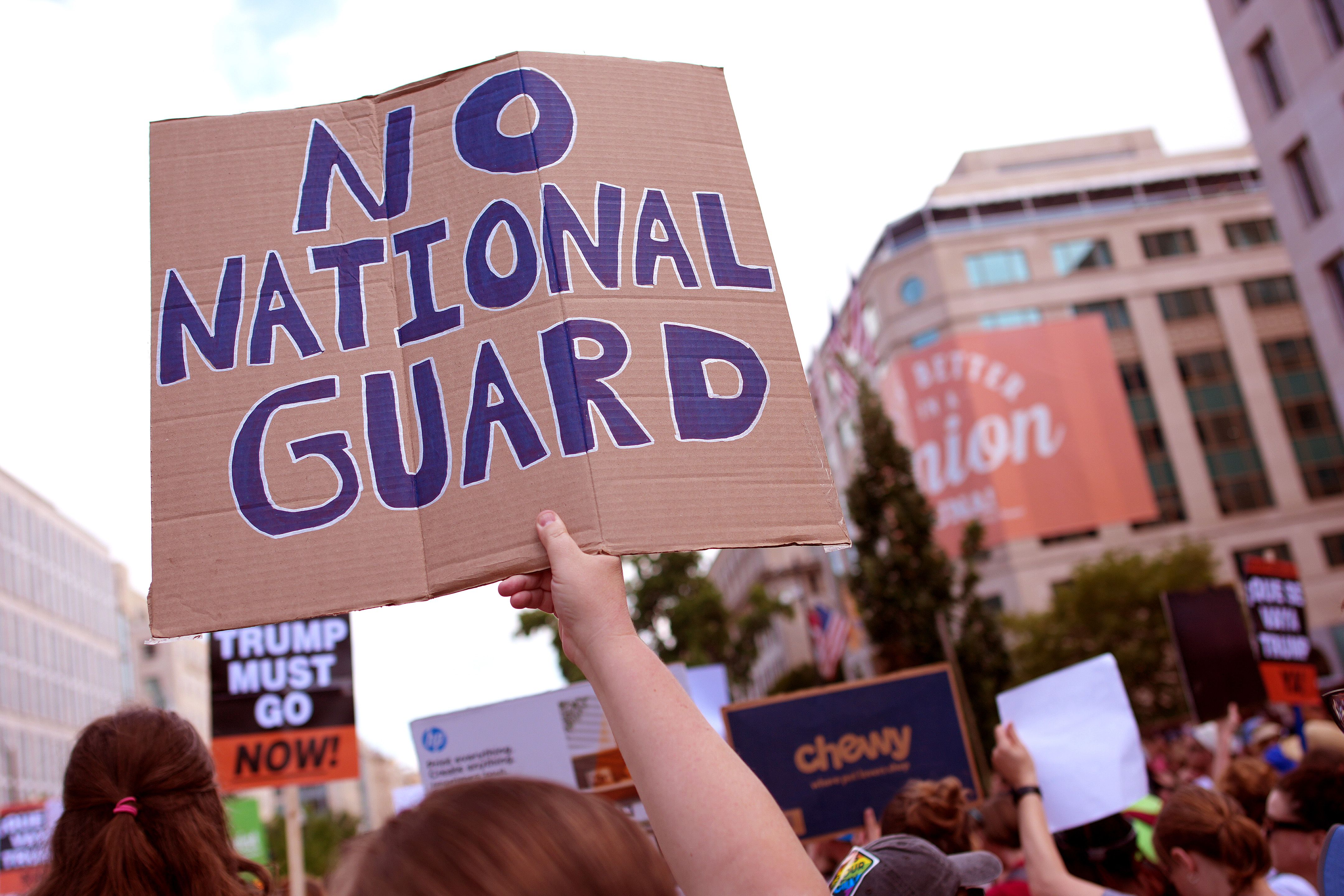 Protesters in a city hold signs, including a large cardboard one in the foreground reading "NO NATIONAL GUARD" in blue letters. Other signs say "TRUMP MUST GO NOW!"