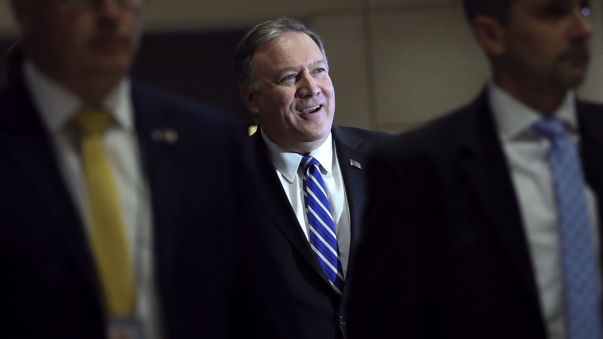 In this image, Secretary of State Mike Pompeo stands in a suit and tie and walks between two men
