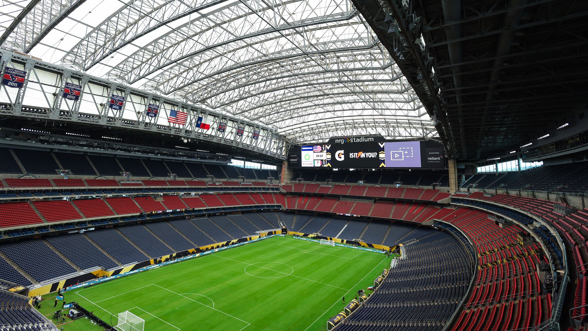 Panoramic view inside a large stadium with a green soccer field, empty red and blue seats, an arched transparent roof, and banners hanging above one side.