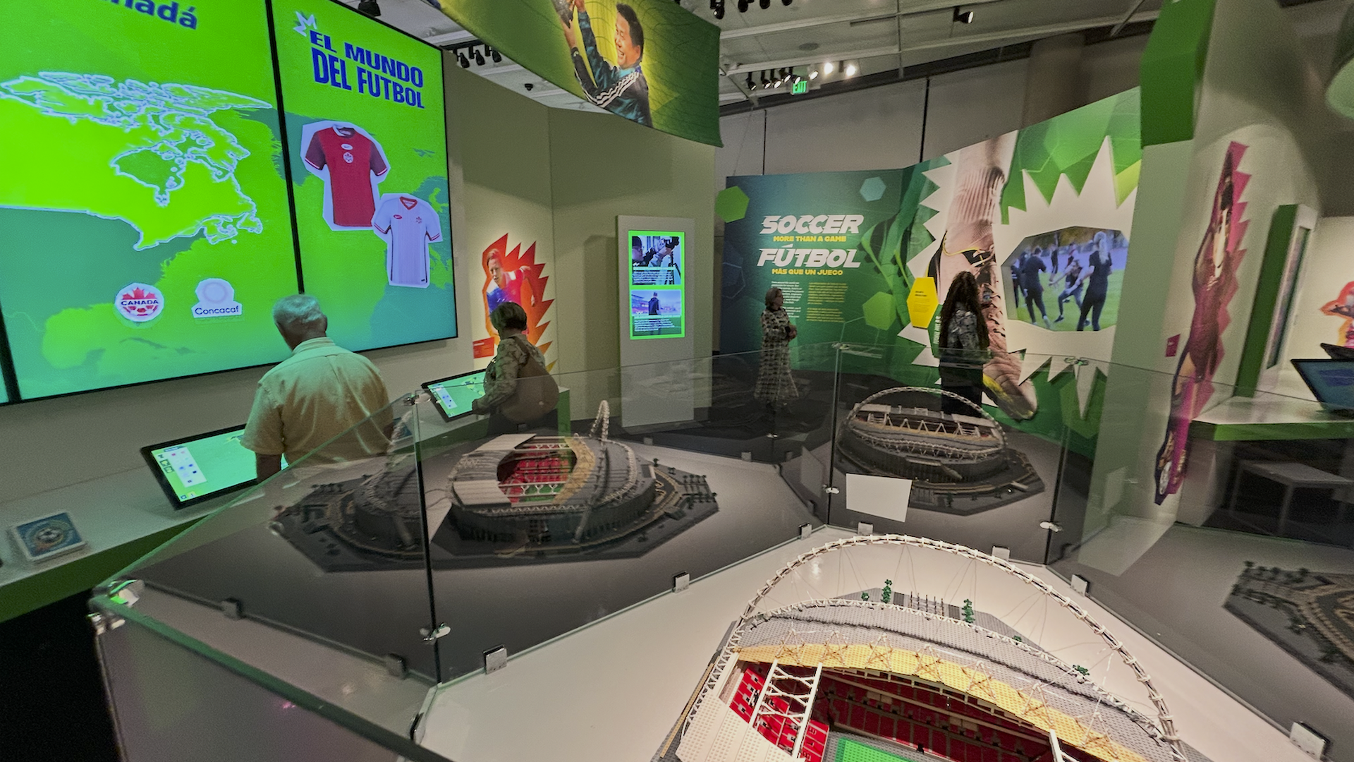 Museum exhibit about soccer with green displays, including Canada map, jerseys, and text "Soccer More Than a Game". People interact with screens. Two stadium models in glass cases.