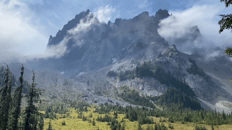 Mountain with sharp rocky peaks partly covered by moving clouds, lush green forest and grassland at the base under a light blue sky.