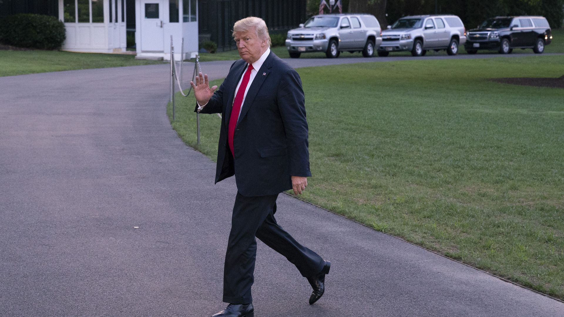 Trump walking across a road on the White House lawn waving to reporters