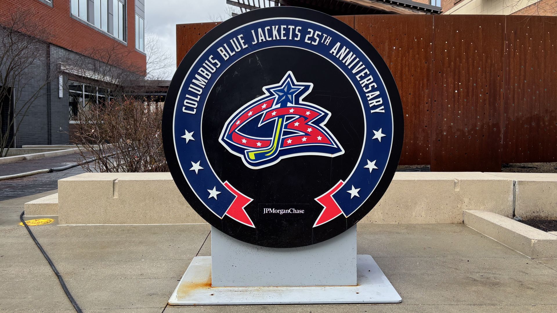 Round black sign with a blue ring reading "Columbus Blue Jackets 25th Anniversary" around a CBJ logo, mounted on a concrete pedestal in an urban plaza with brick buildings nearby; JPMorgan Chase at bottom.