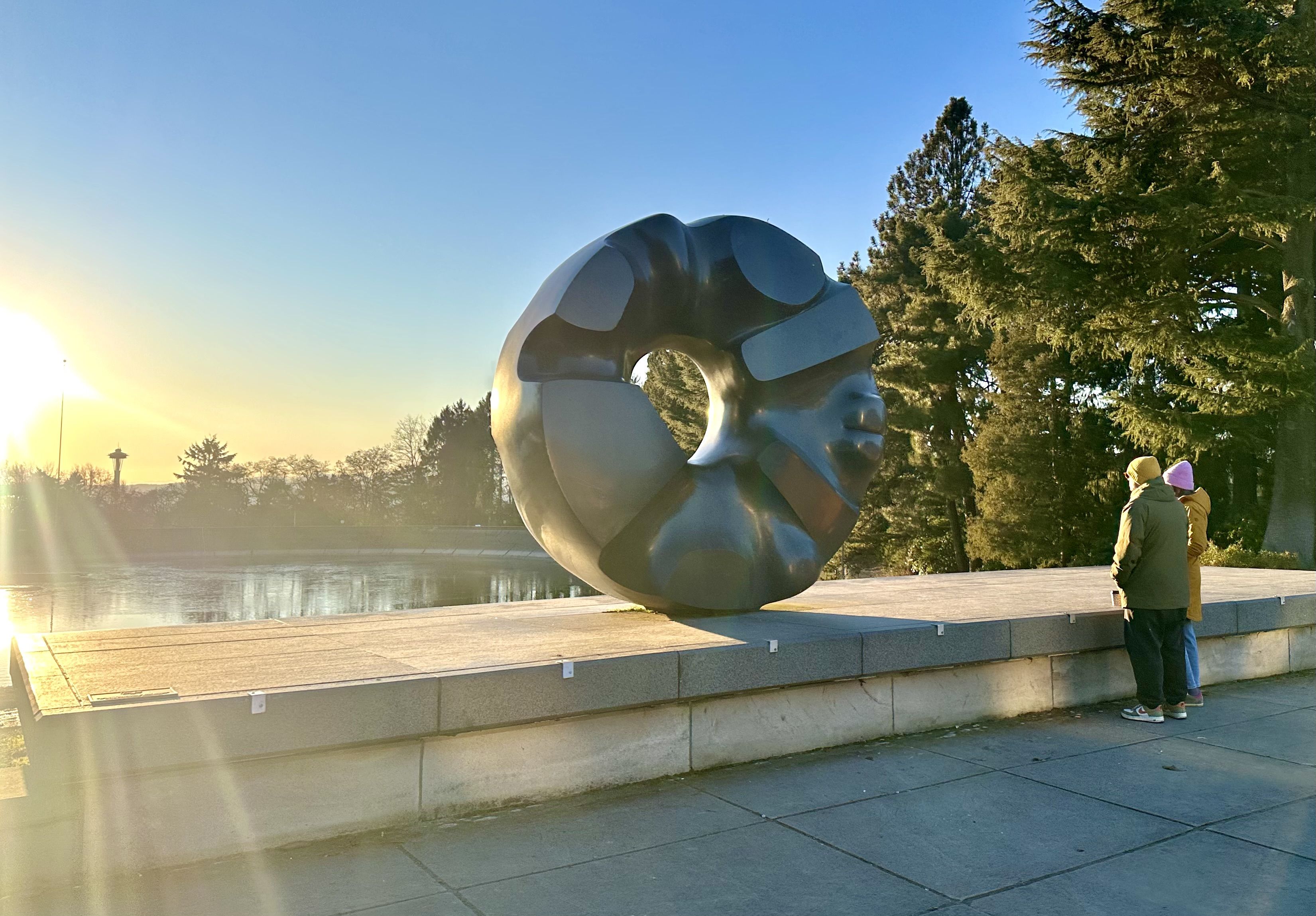 Two people look out at at the setting sun by the Black Sun sculpture at Volunteer Park, with trees in the background and reflections of the sun off a reservoir of water.