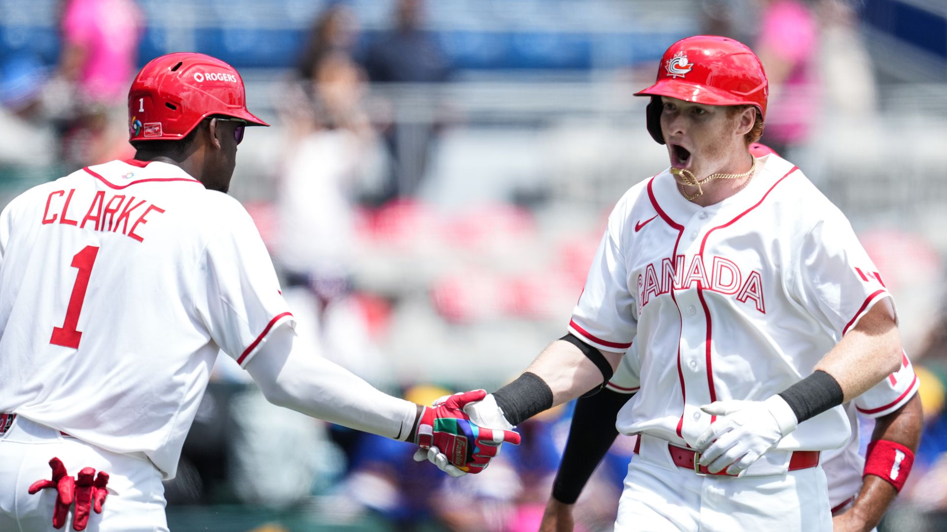 Two basebal players wearing white Team Canada jersies shake hands in celebration and one of them runs the bases, yelling.