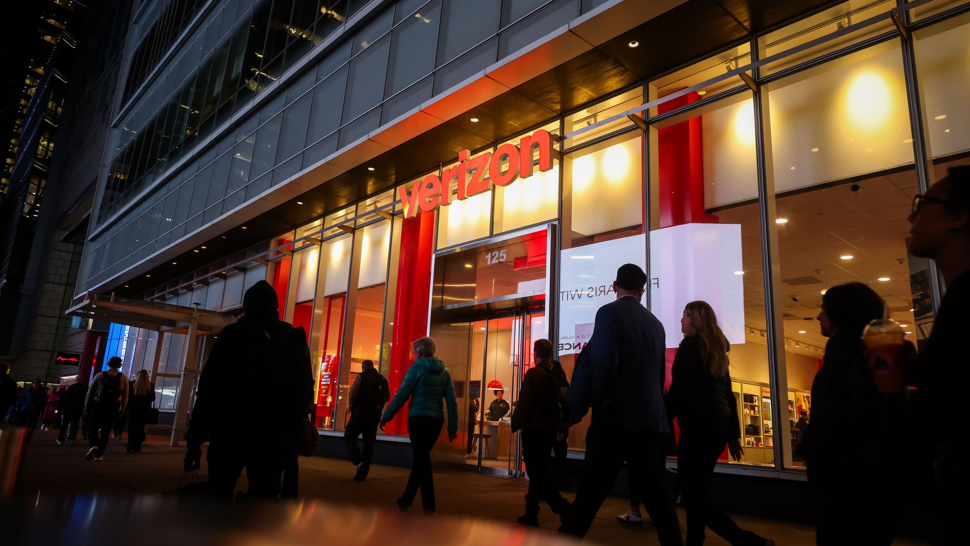 Evening street scene with people walking past an illuminated Verizon store with large glass windows and a red and white interior in an urban area.