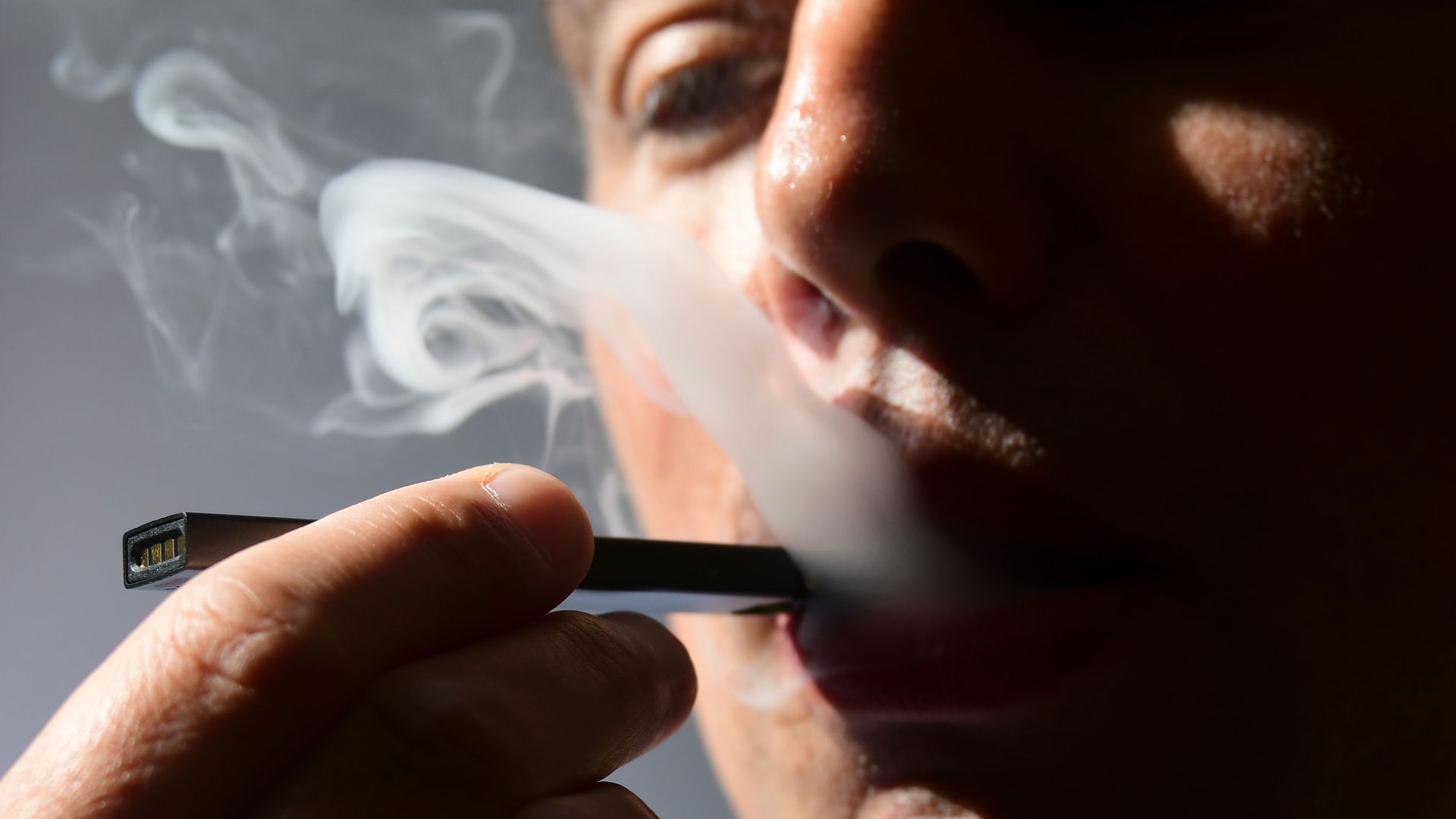 In this image, a person holds a vape pen to their mouth and blows smoke at the same time;