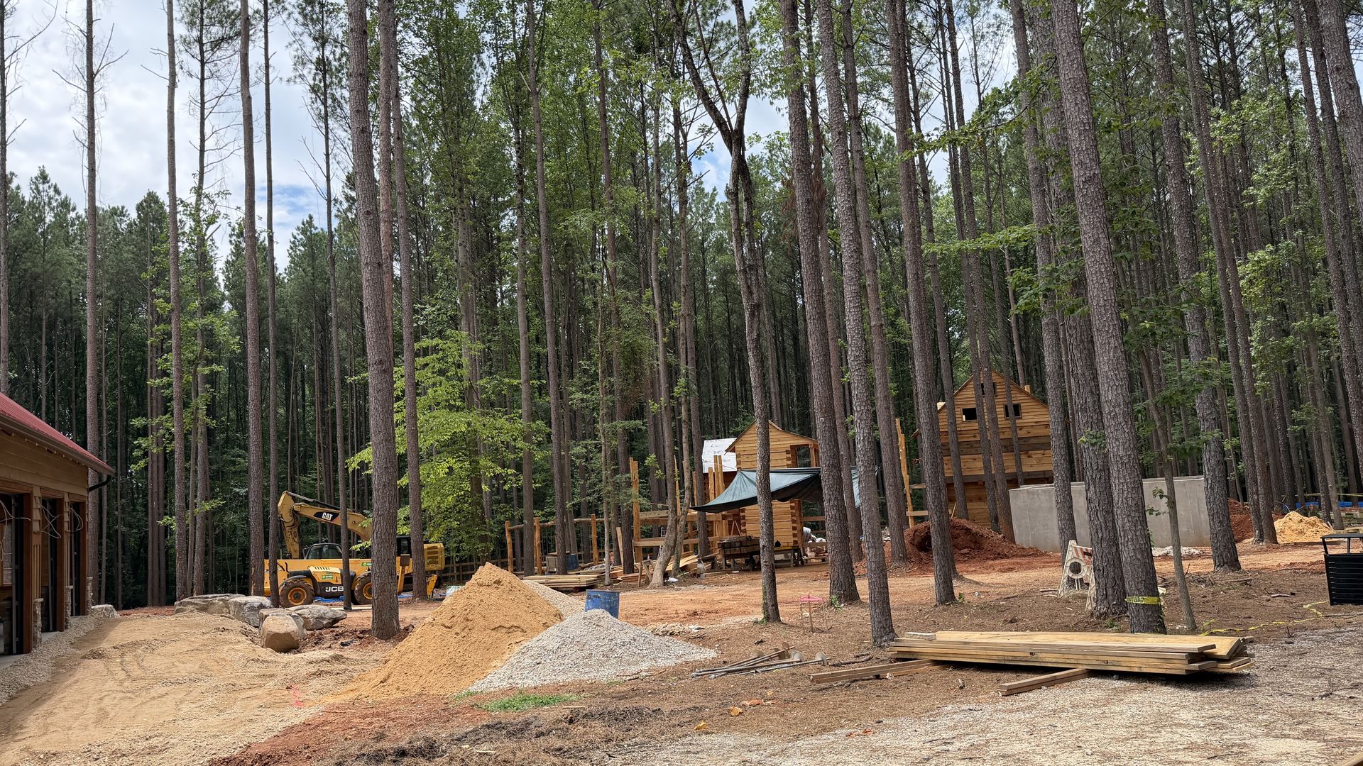 Panoramic view of a forested construction site. Tall pine trees surround partially built wooden cabins, orange dirt piles, gravel, and a small excavator at work in the foreground.