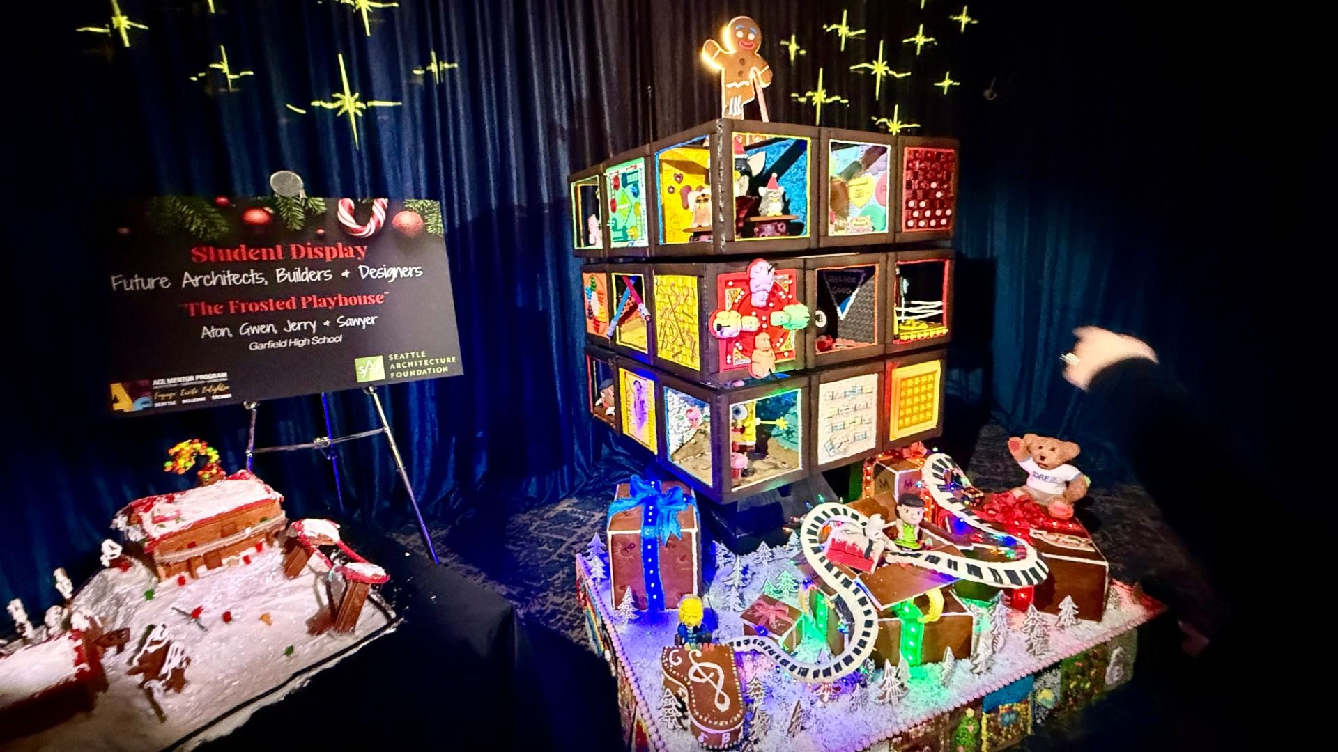Colorful gingerbread display with decorated cubes, musical notes, a gingerbread man, and a bear, set against dark blue backdrop with star lights. Another gingerbread display titled The Frosted Playhouse is at left.