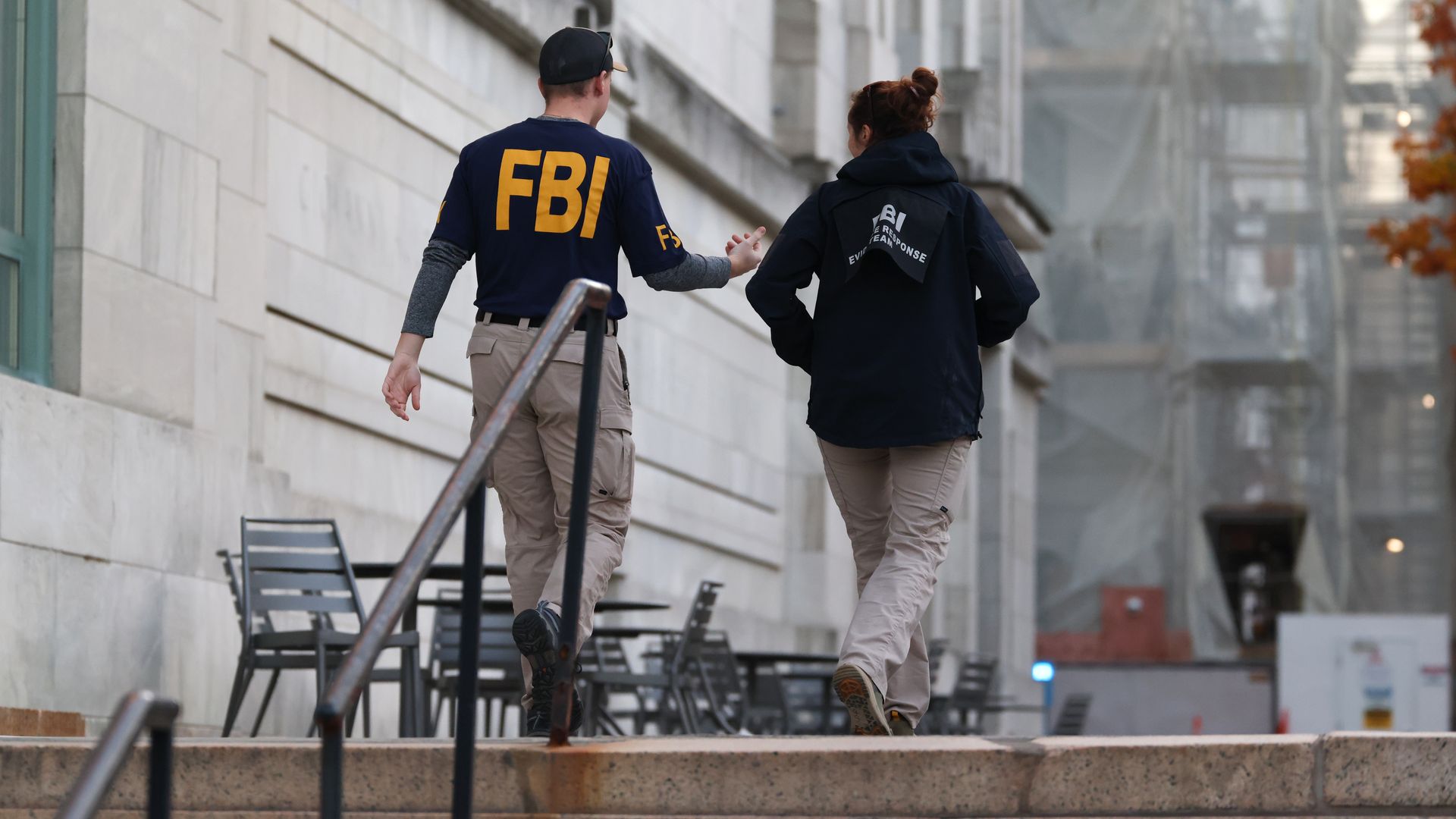 Two FBI agents in beige pants and dark jackets with yellow FBI letters walk up outdoor stone steps beside a light stone building with outdoor chairs and a railing.