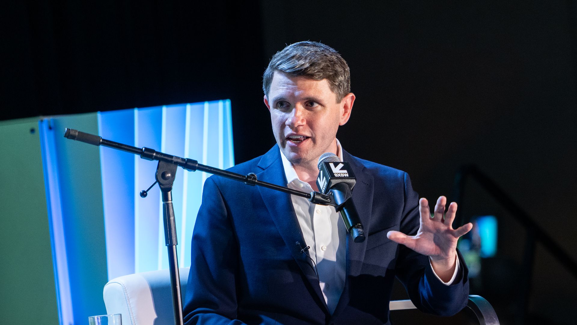 Man in a navy suit and white shirt sits on a stage, speaking into a mic on a stand. He gestures with his right hand; blue lighting and a glass of water sit nearby.