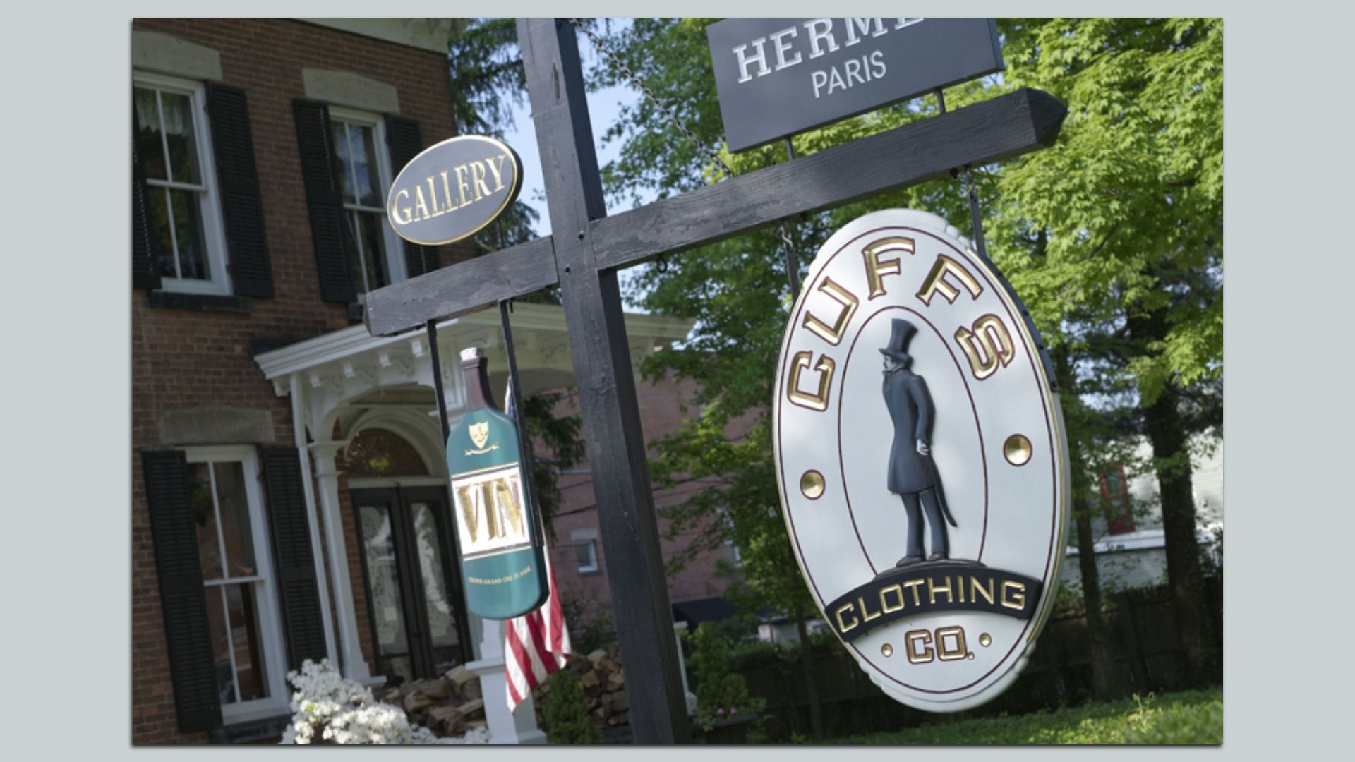 Outdoor signs in front of a brick building with black shutters: "GALLERY," a green wine bottle, "HERMES PARIS," and a white oval sign with a man in a top hat for "CLUFFS CLOTHING CO."