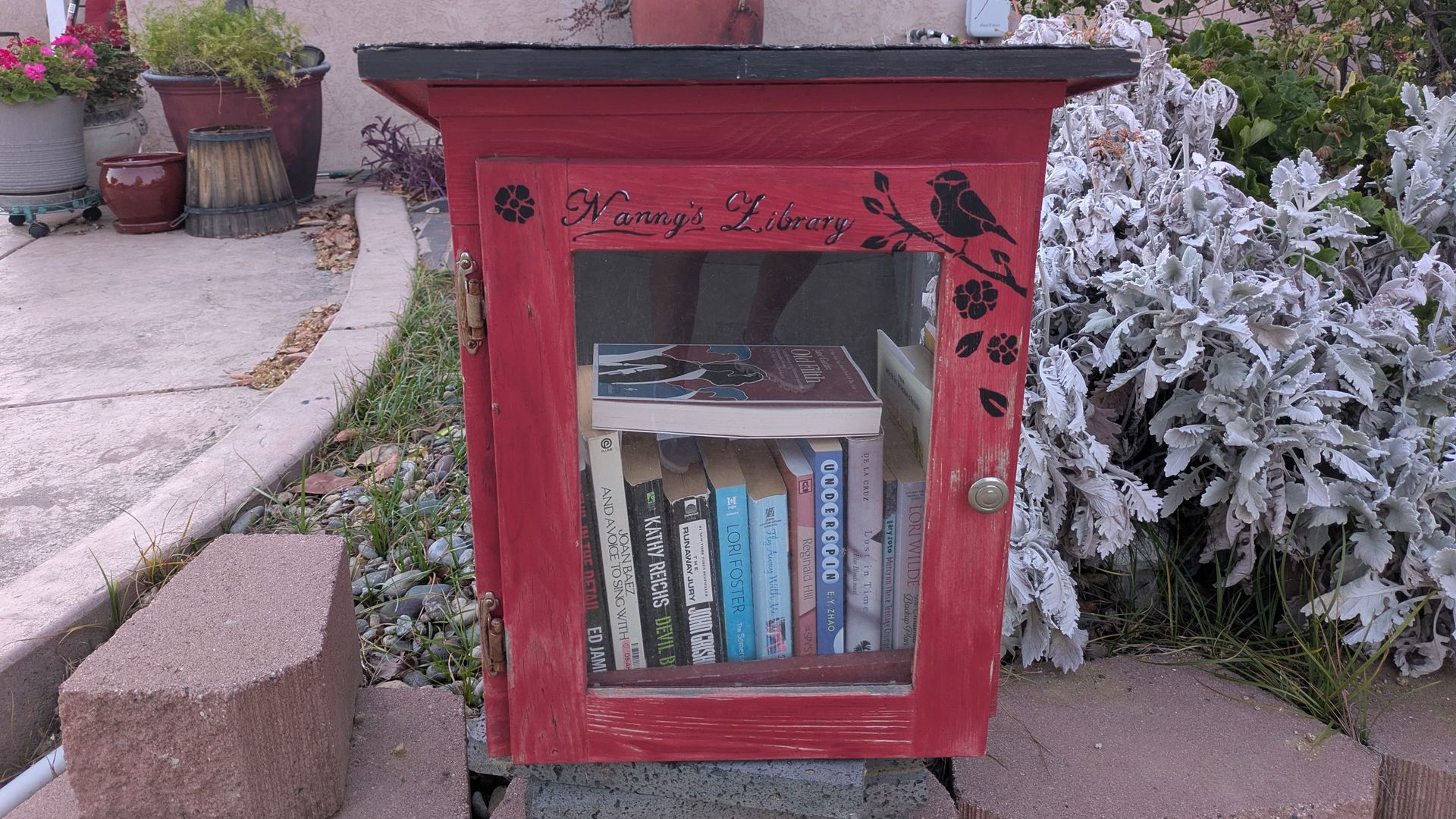 Outdoors, a red wooden library box labeled "Nanny's Library" holds books; a large red vase sits on top. Surrounded by potted plants and silvery foliage near a white house with a window.