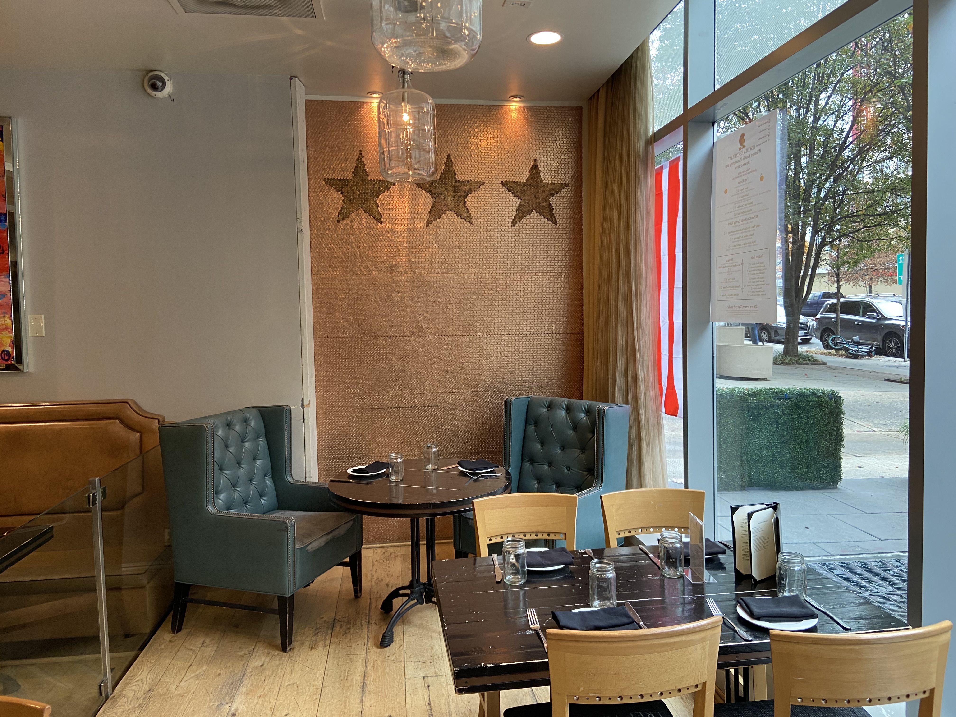 Cozy restaurant corner with blue tufted chairs around a round table and wooden chairs at a rectangular table, set with black napkins and mason jars, next to large windows showing a sidewalk.