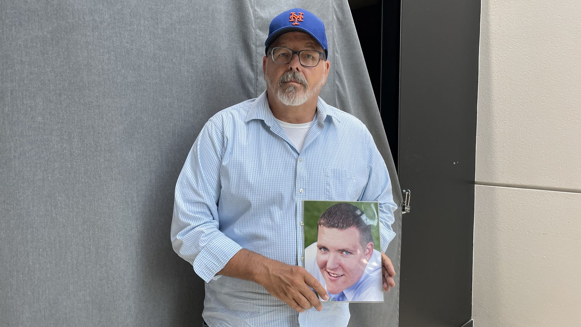 A man in a blue shirt and blue baseball cap holds a photo of his son. 