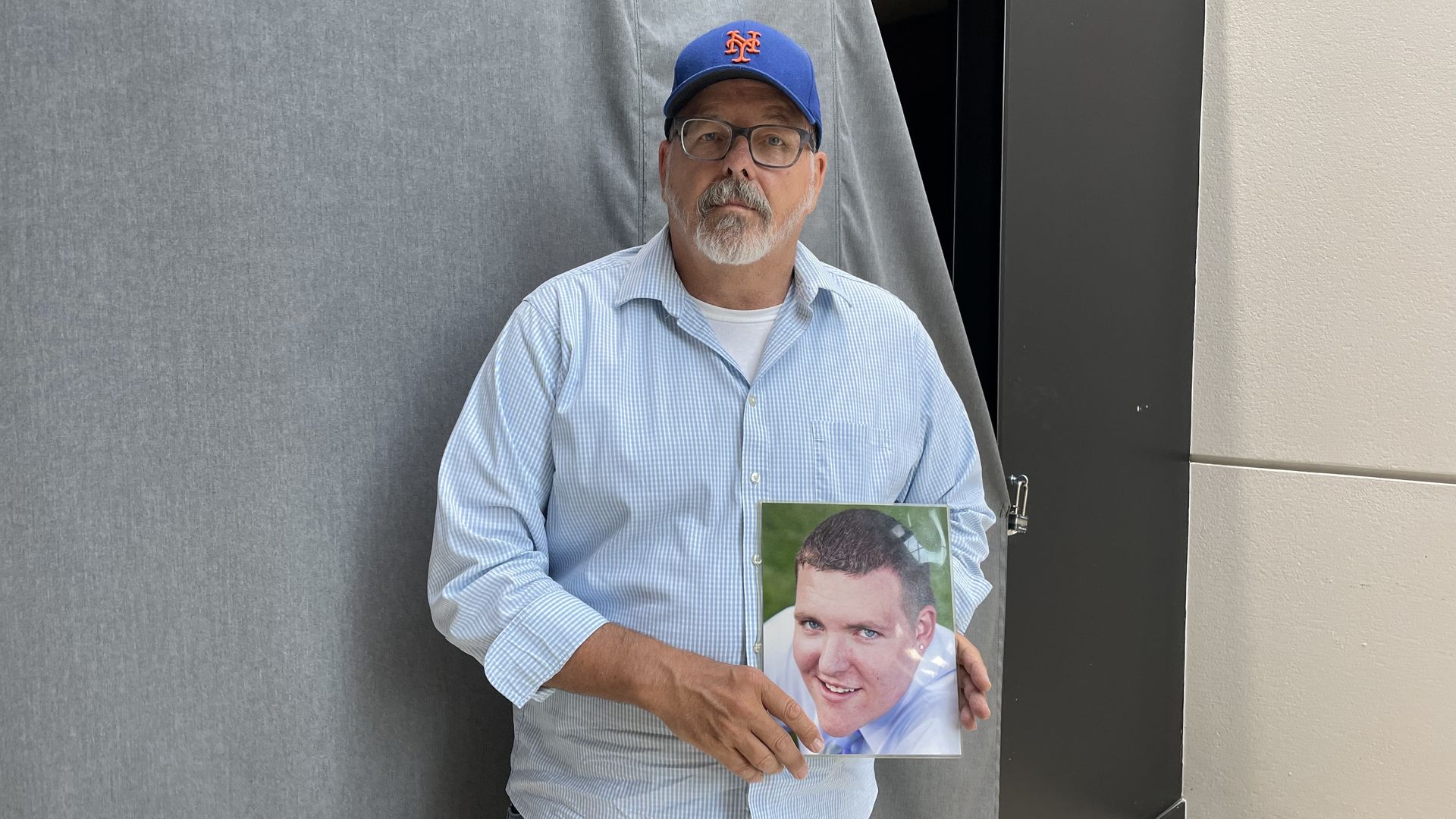 A man in a blue shirt and blue baseball cap holds a photo of his son.