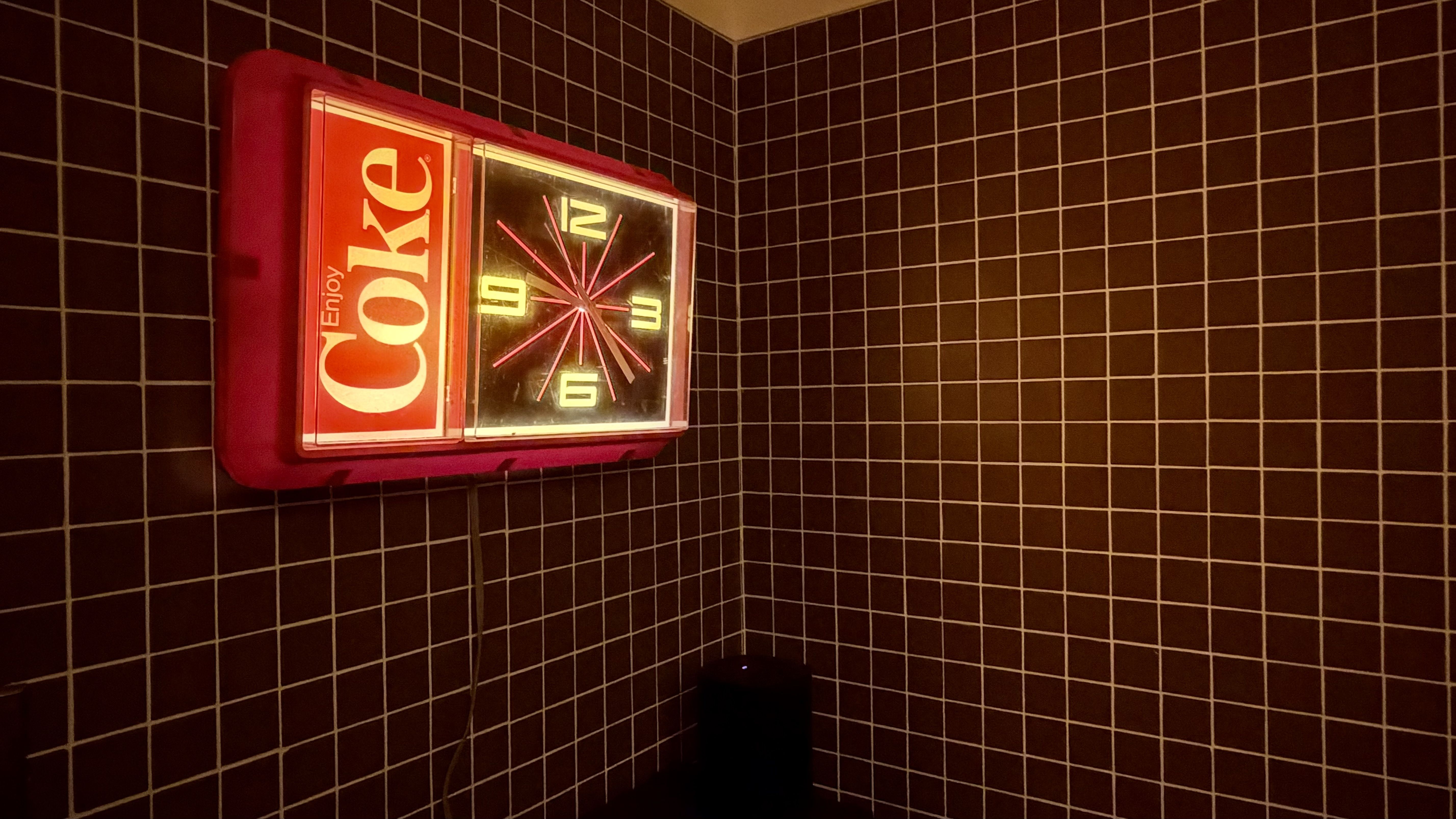 Red Coca-Cola clock with neon pink starburst clock face, left panel shows "Coke" and "Enjoy" text, mounted on a dark brown tiled wall in a corner; warm glow, and a dark object on the floor.