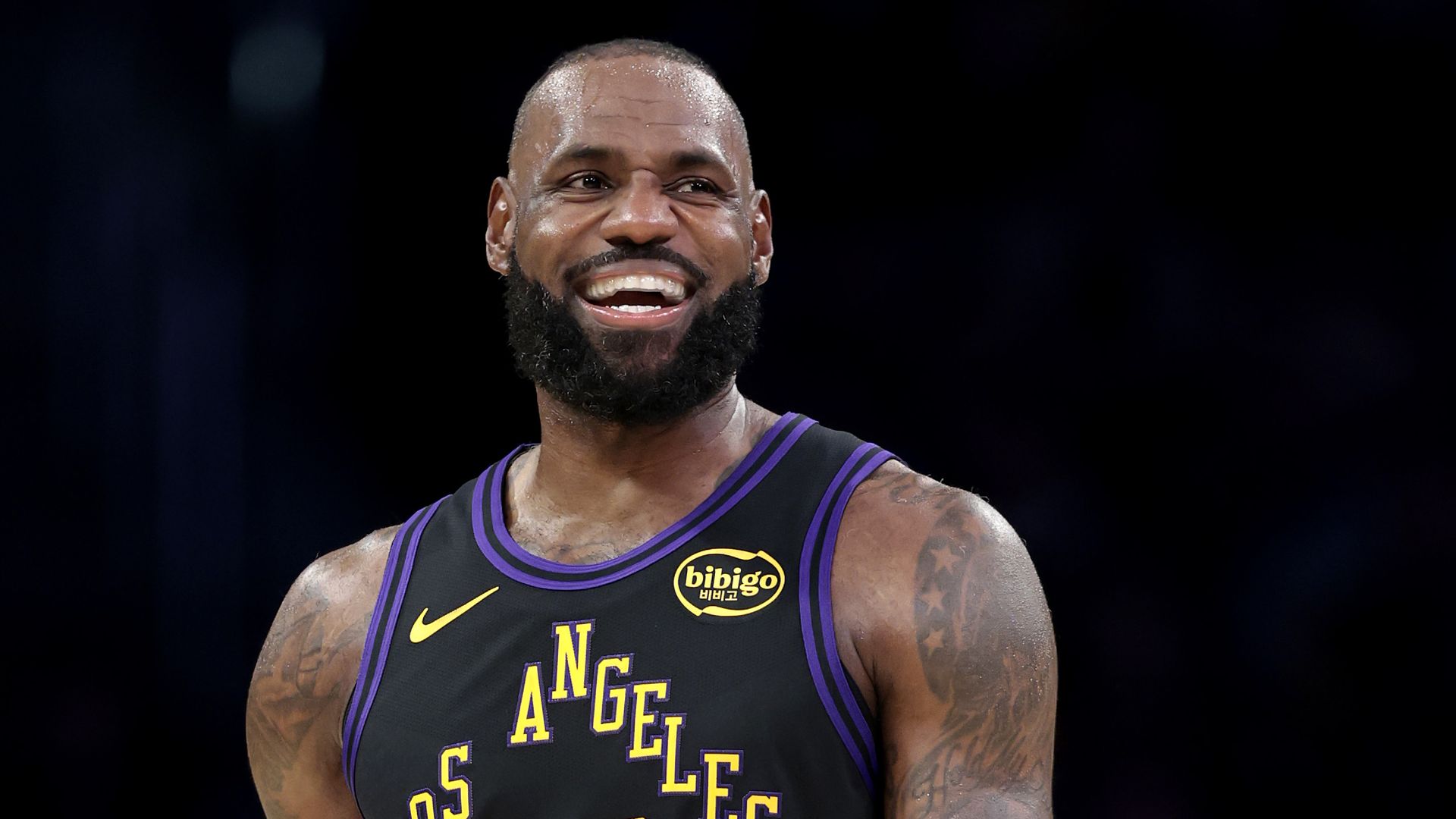 Close-up of a smiling male basketball player with a full beard and short hair, wearing a black Lakers jersey with purple and gold trim and the number 23.