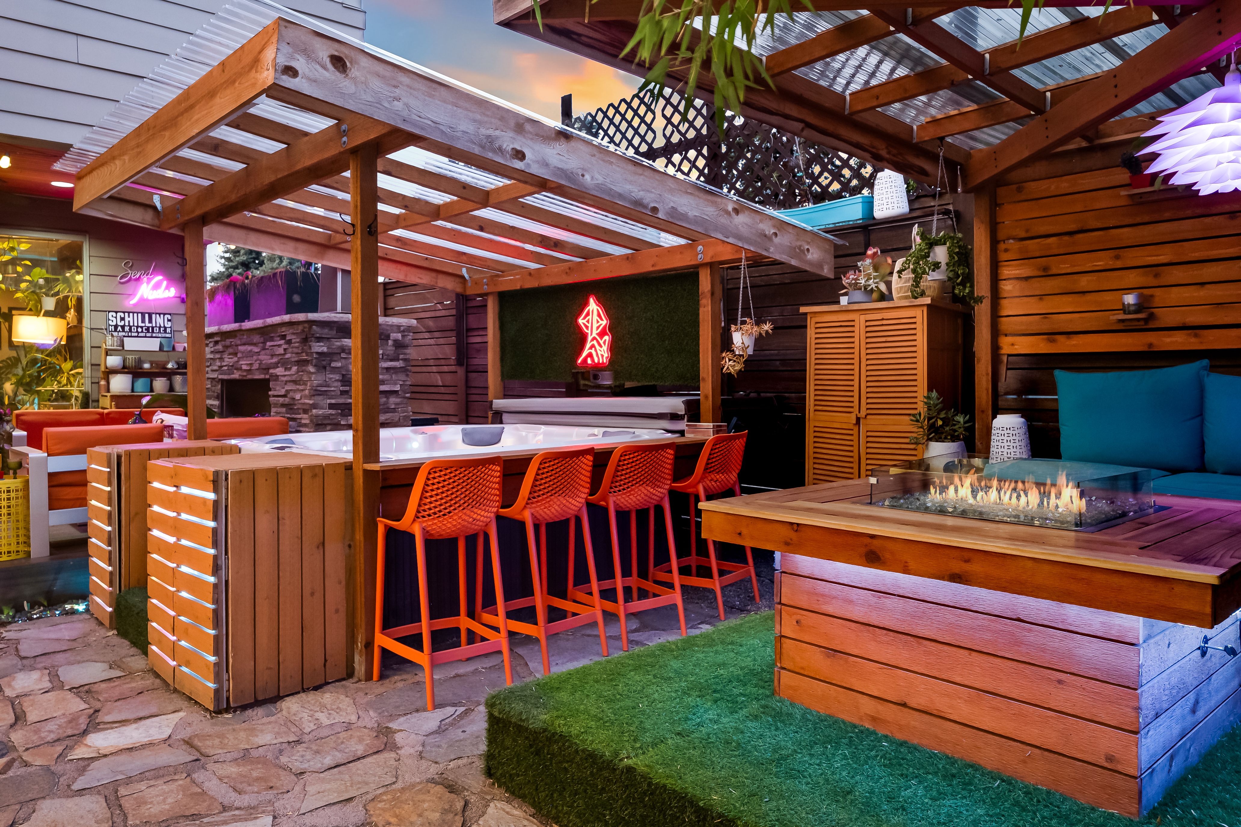 Outdoor patio with hot tub,  orange bar stools, a stone fireplace, a fire pit table on artificial grass, blue cushions, hanging plants and neon signs at dusk.