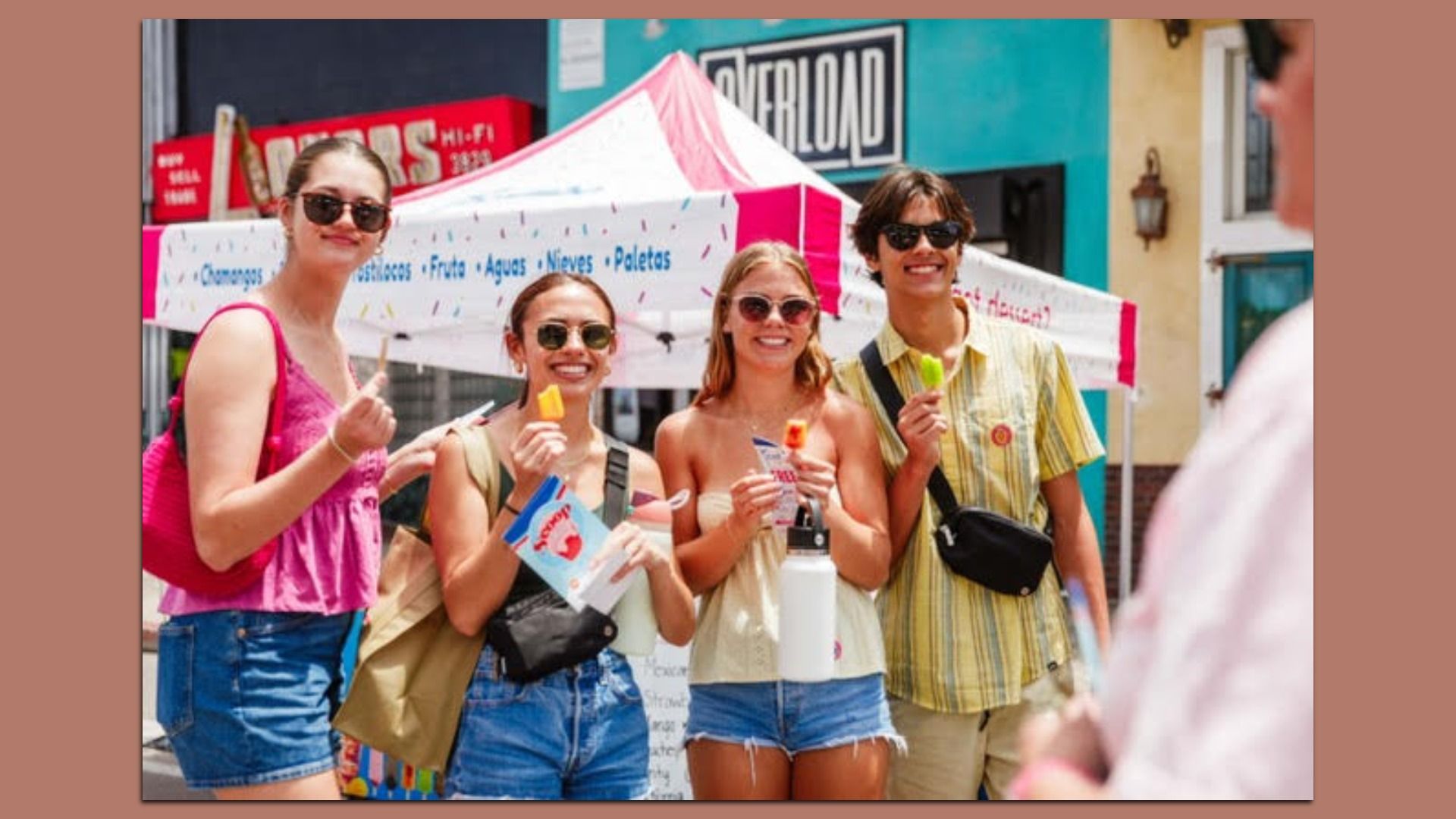 Four friends wearing sunglasses smile and hold colorful ice pops at a sunny street festival, standing in front of a white-and-pink tent with a teal building in the background.