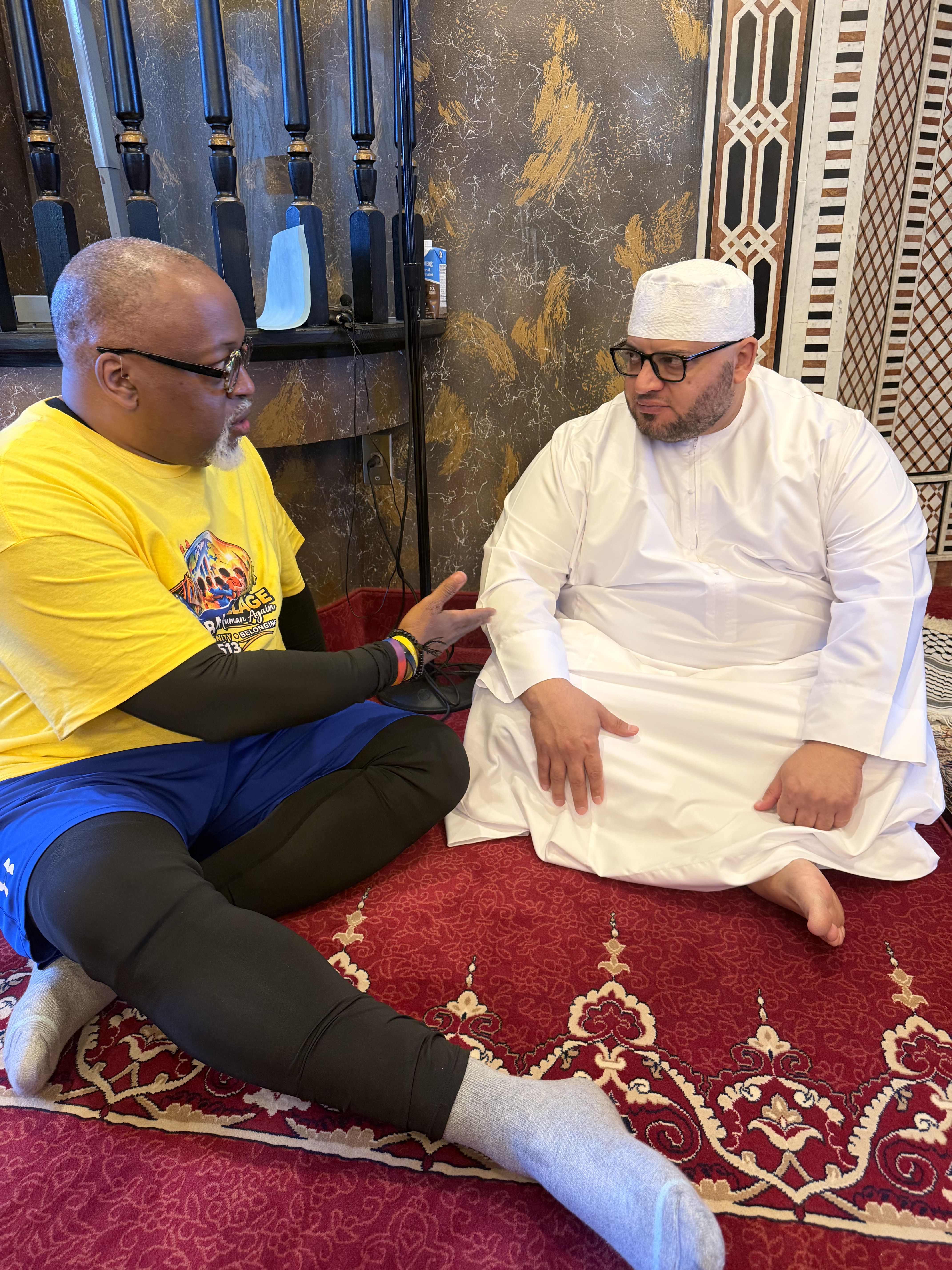 Two men sit on a red patterned carpet facing each other and talking. Left: yellow T‑shirt; right: white traditional outfit with a kufi. Ornate marble wall with geometric panels behind; a blue mask rests on the railing.