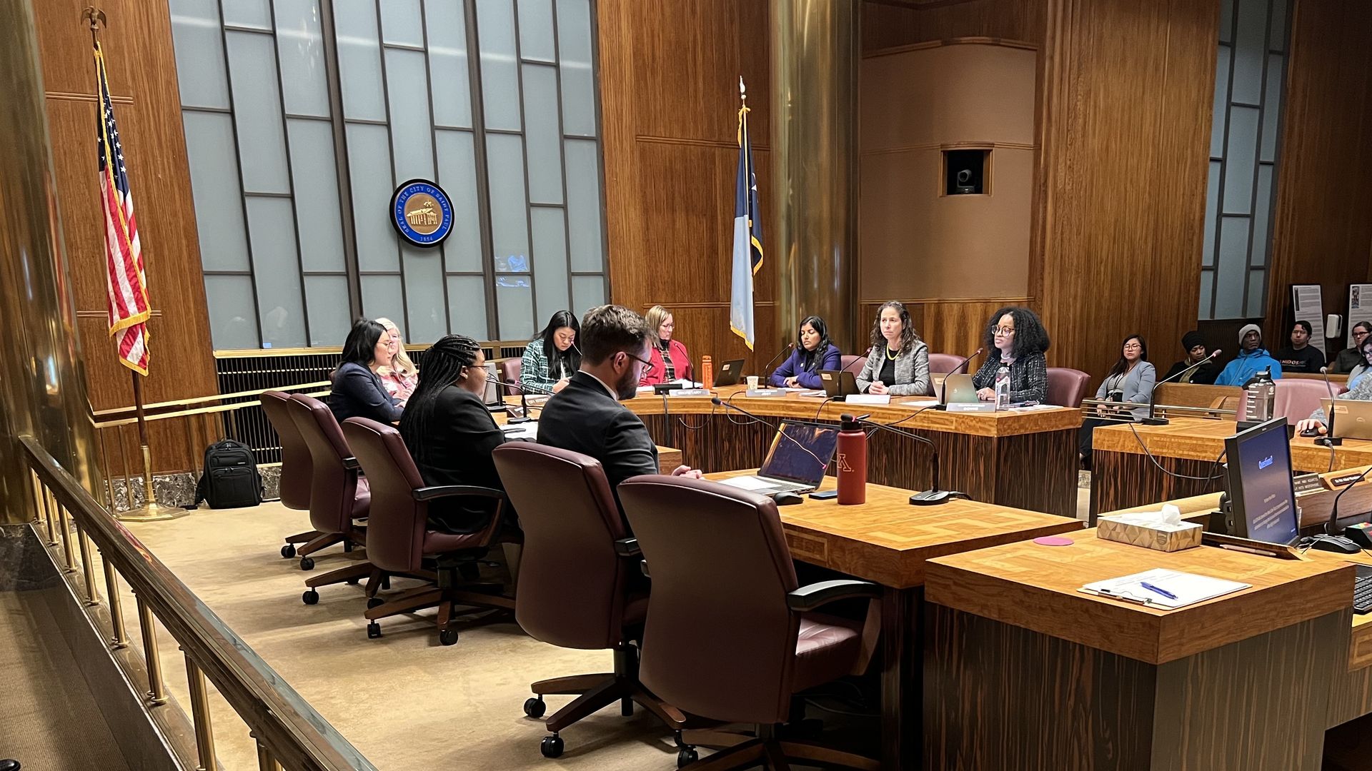 A city council chamber during a meeting with several people and staff sitting around a horseshoe-shaped dais