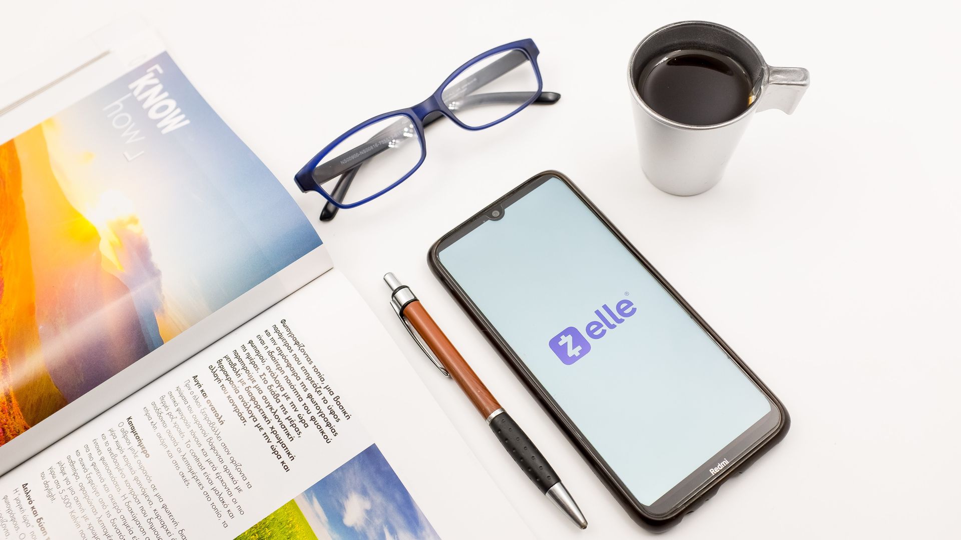 Smartphone with Zelle logo next to a pen, open textbook, glasses, and a thermos with coffee