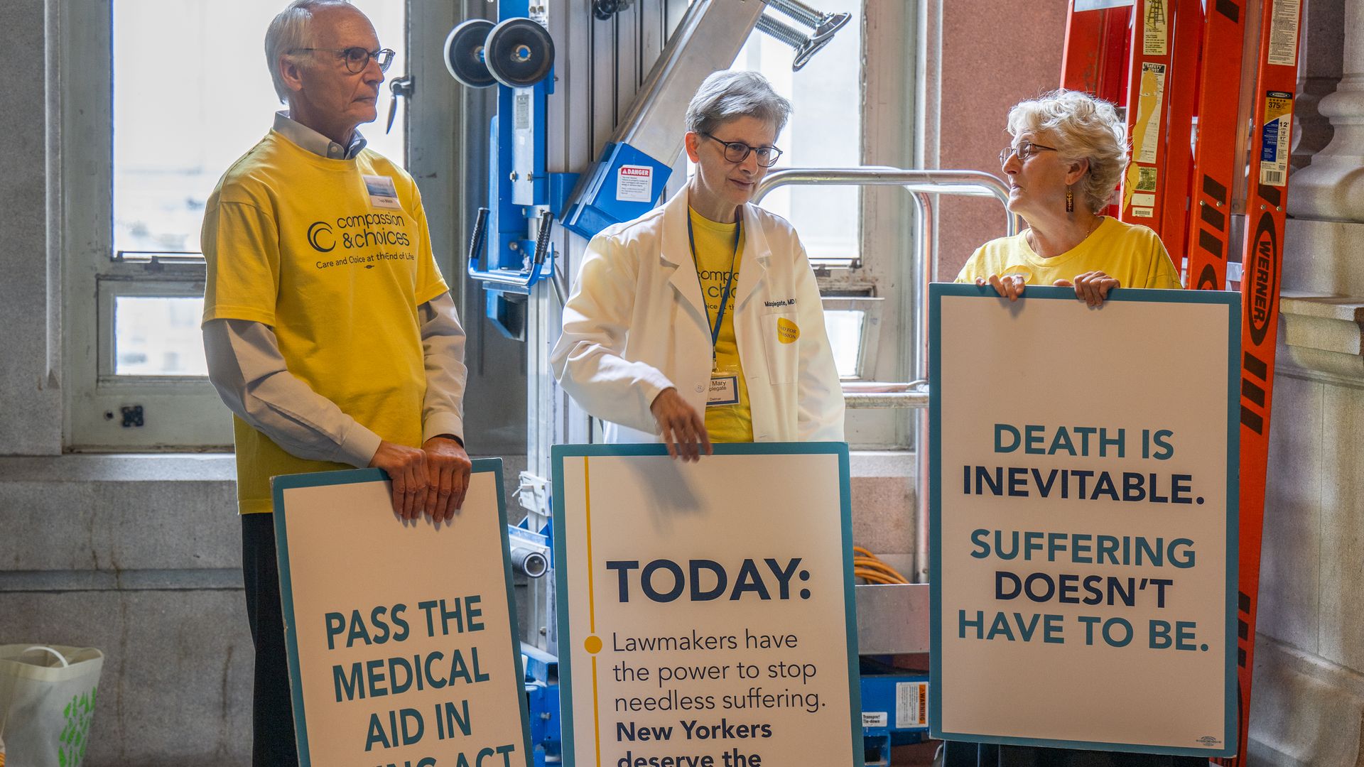Three people in yellow shirts stand in an industrial-looking space, one wearing a white lab coat, holding large protest signs about death and medical aid; orange safety equipment visible behind.