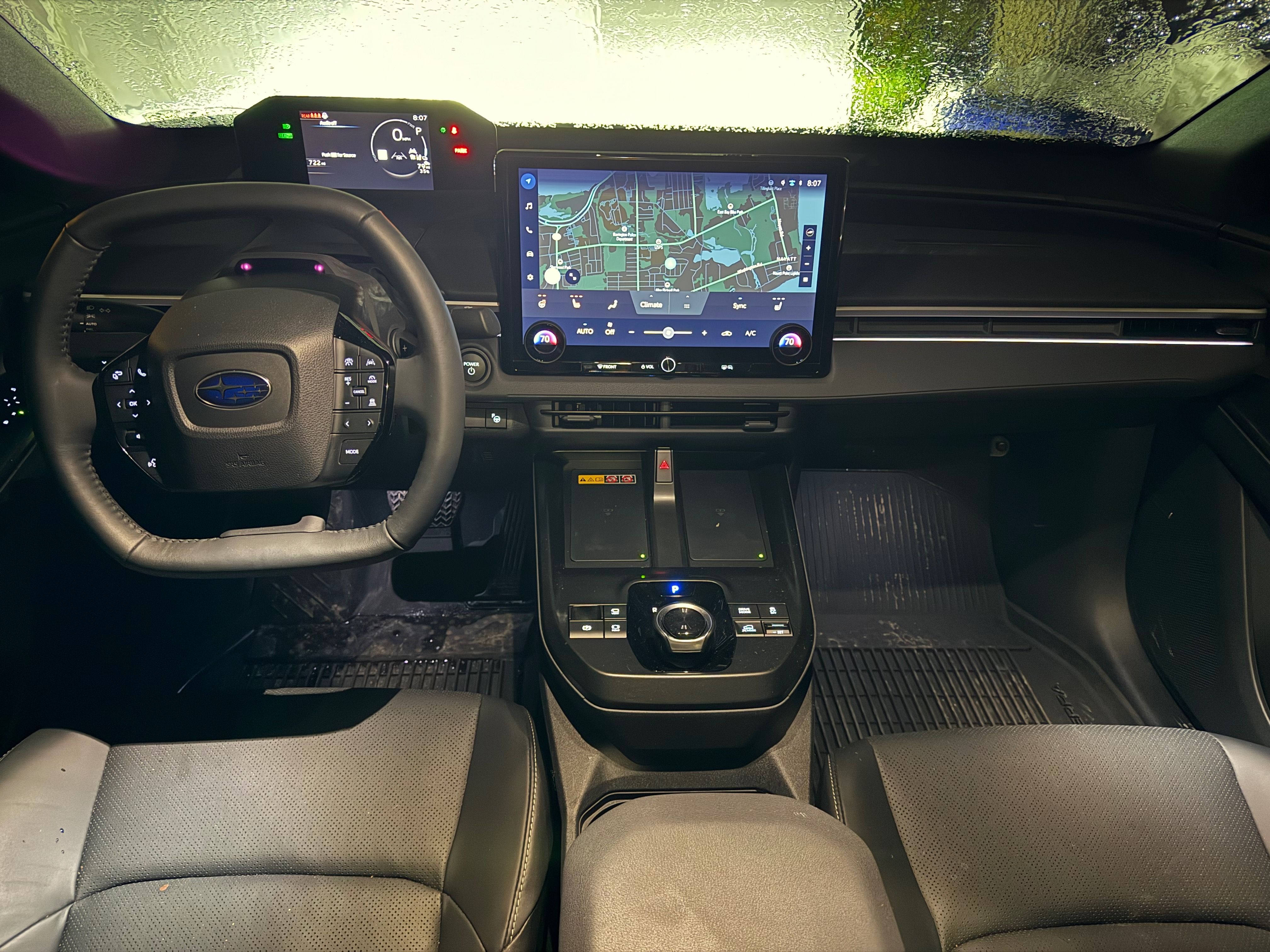 Car interior with a left-hand steering wheel featuring the Subaru logo, a large touchscreen showing navigation, climate controls below, and a rotary gear selector on a black center console; dark seats.
